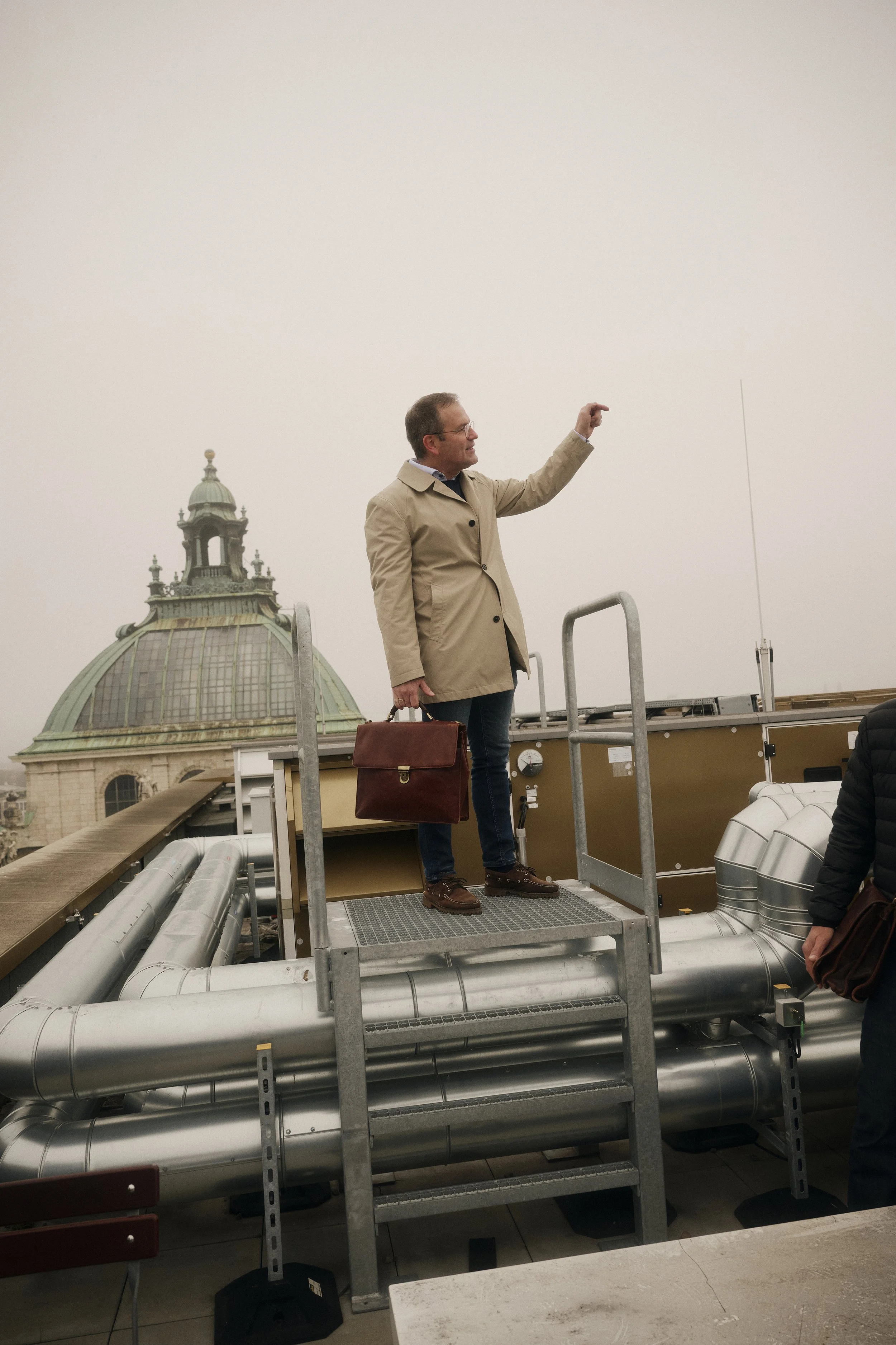 Eine Person in einem beigen Mantel steht auf einem metallenen Podest auf einem Dach mit metallischen Rohren. Im Hintergrund ist die Kuppel eines historischen Gebäudes zu sehen. Die Person hält eine Aktentasche und zeigt mit der Hand in die Ferne.