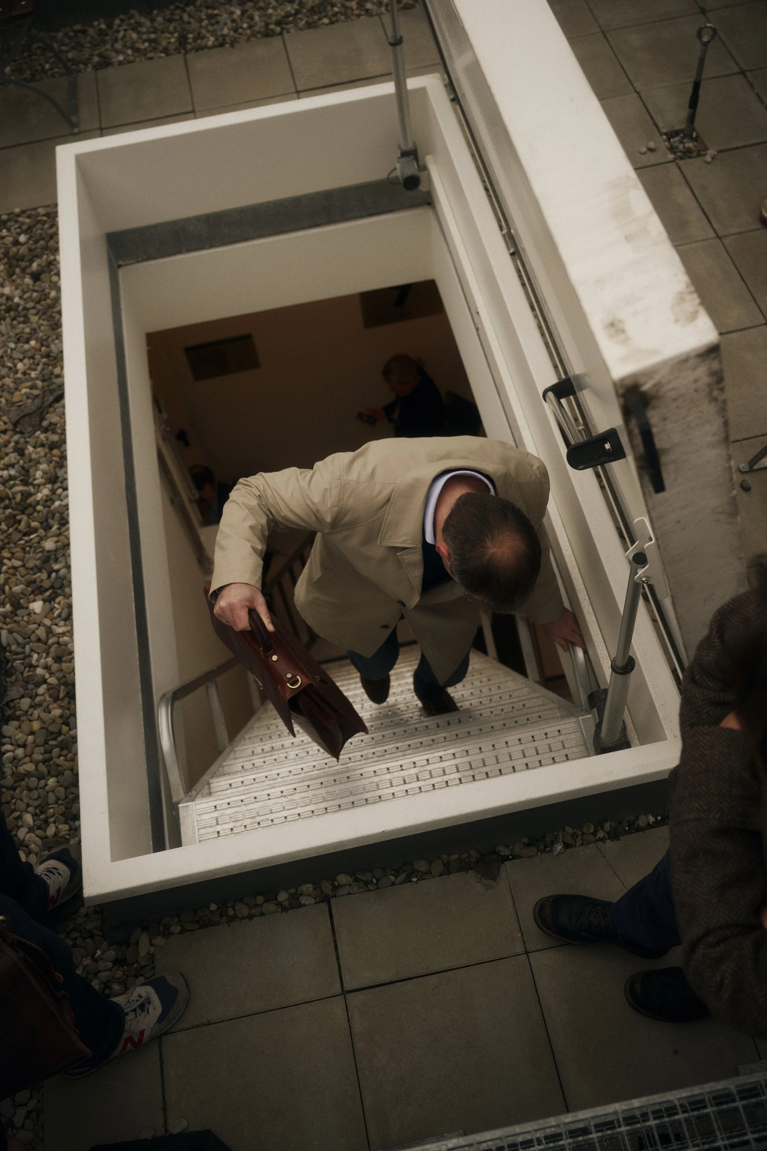 Ein Mann geht eine Treppe hinauf, trägt einen beigen Mantel und hält eine braune Ledertasche. Die Treppe führt zu einem Innenraum, und es befinden sich weitere Personen im Hintergrund. Der Boden um die Treppe ist mit Fliesen und Kieselsteinen bedeckt