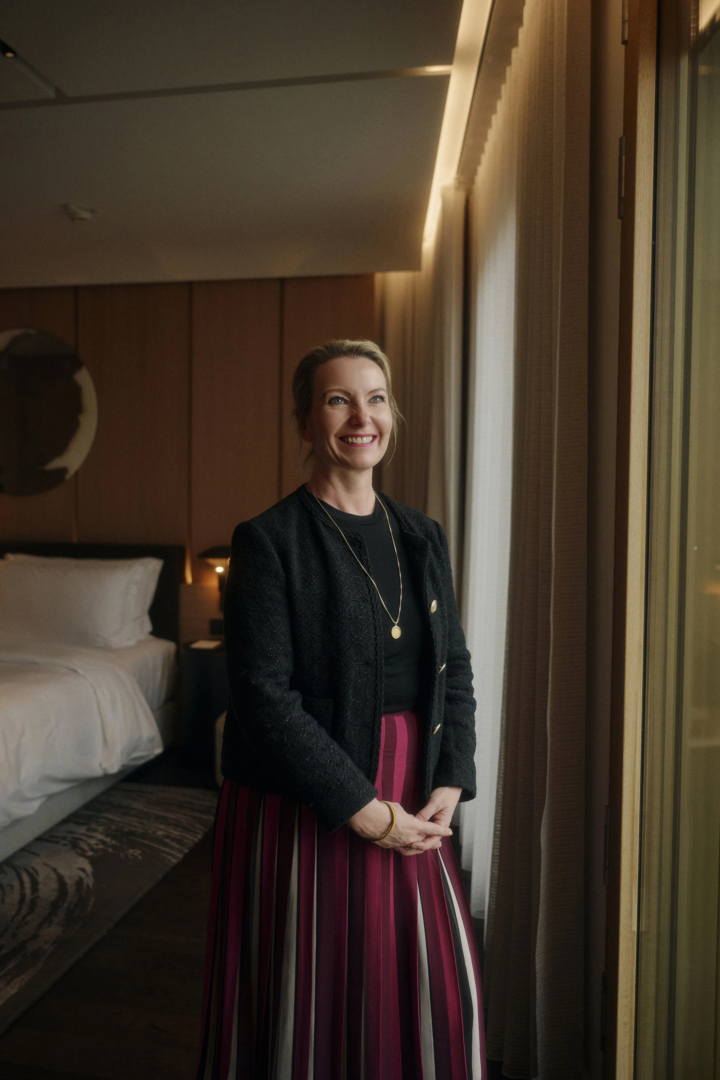 Eine lächelnde Frau steht in einem stilvollen Raum vor einem Fenster. Sie trägt einen schwarzen Blazer und einen gestreiften Rock. Im Hintergrund ist ein Bett mit weißen Kissen zu sehen.