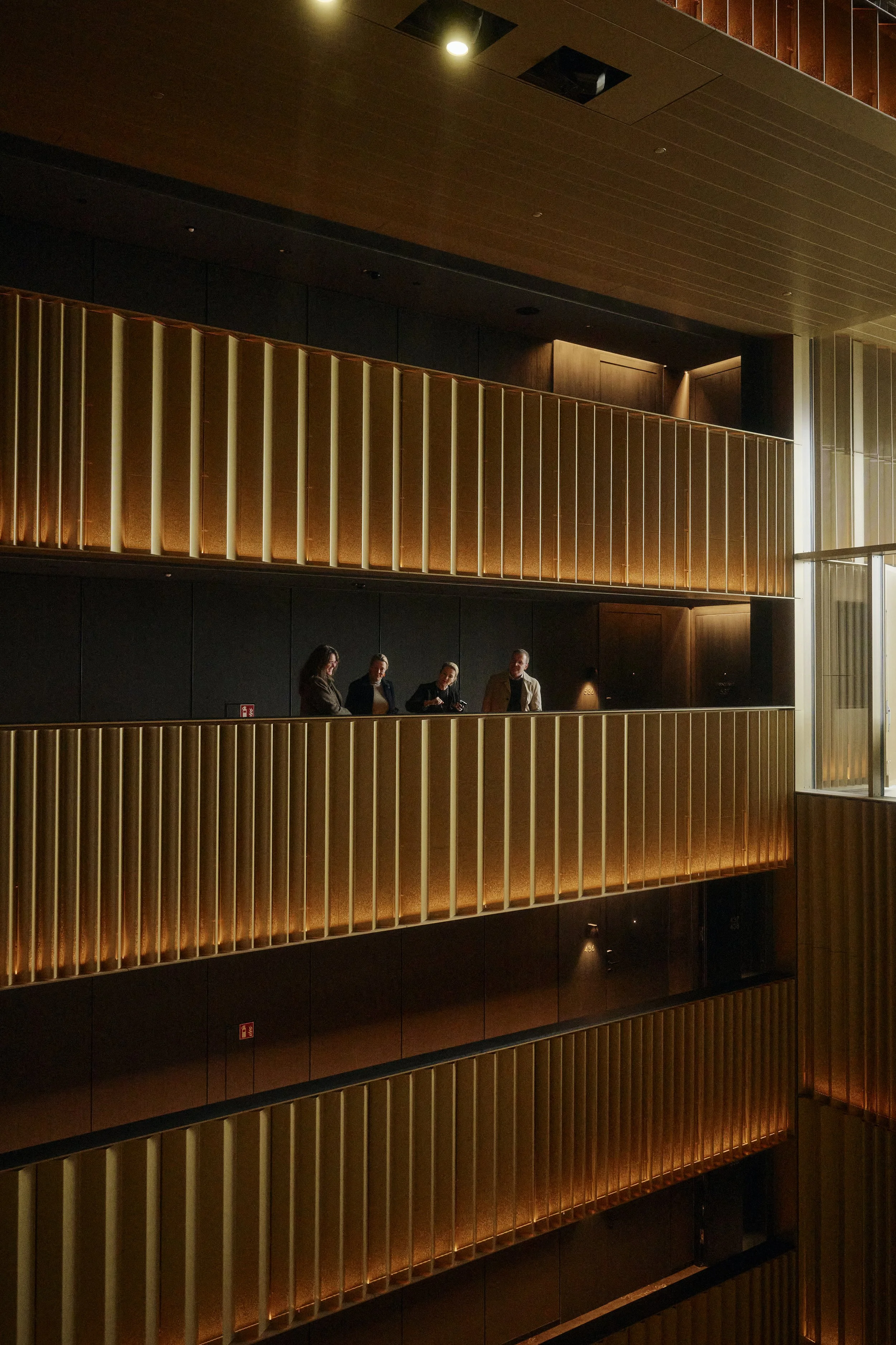 Innenraum eines modernen Gebäudes mit mehreren Etagen und beleuchteten Balkonen, auf denen vier Menschen stehen. Die Gestaltung ist modern mit vertikalen Lamellen und warmer Beleuchtung.