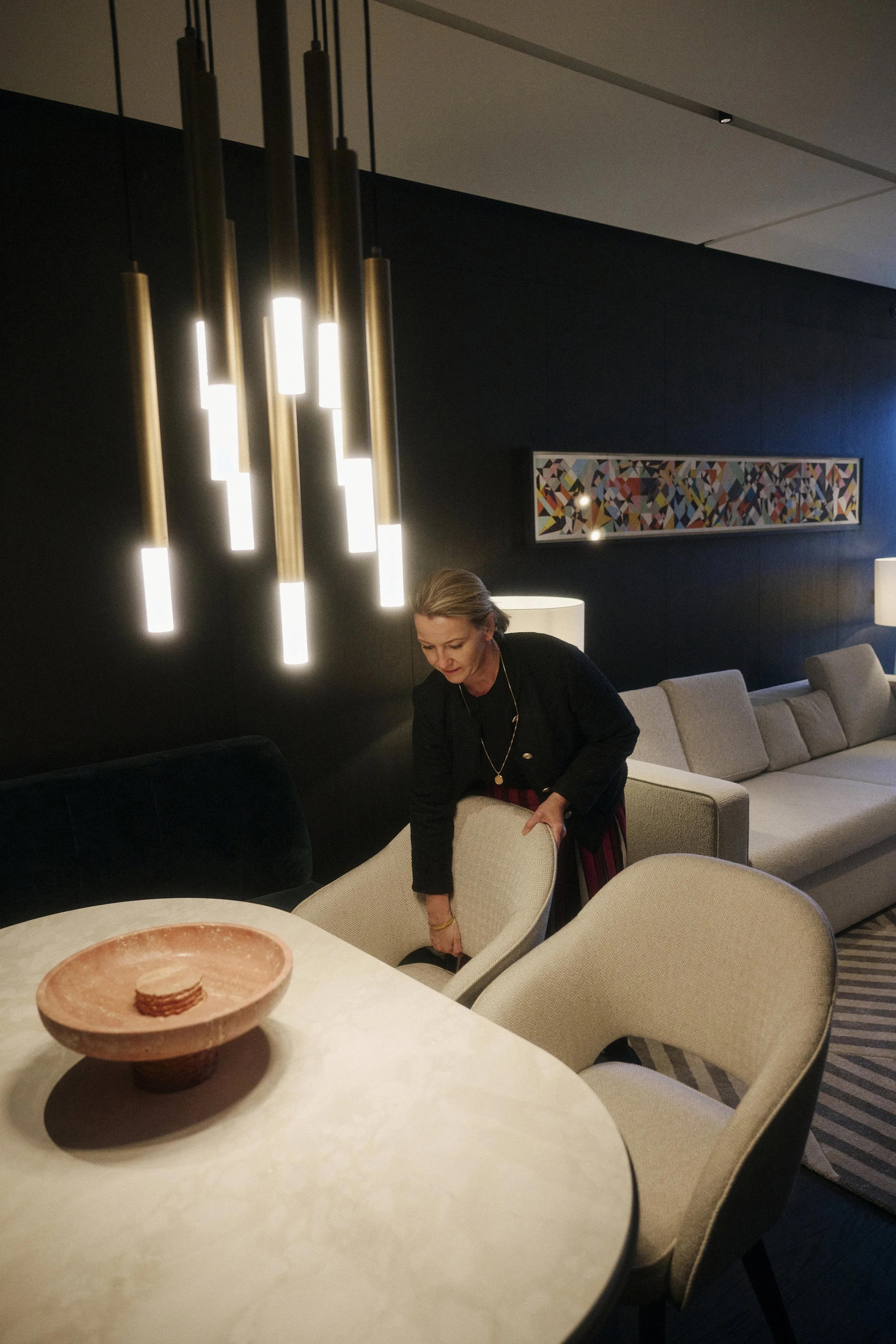 Eine Frau richtet einen Stuhl in einem modern eingerichteten Wohnzimmer mit Designerleuchten, einem Tisch mit Schale und einem Gemälde an der Wand.