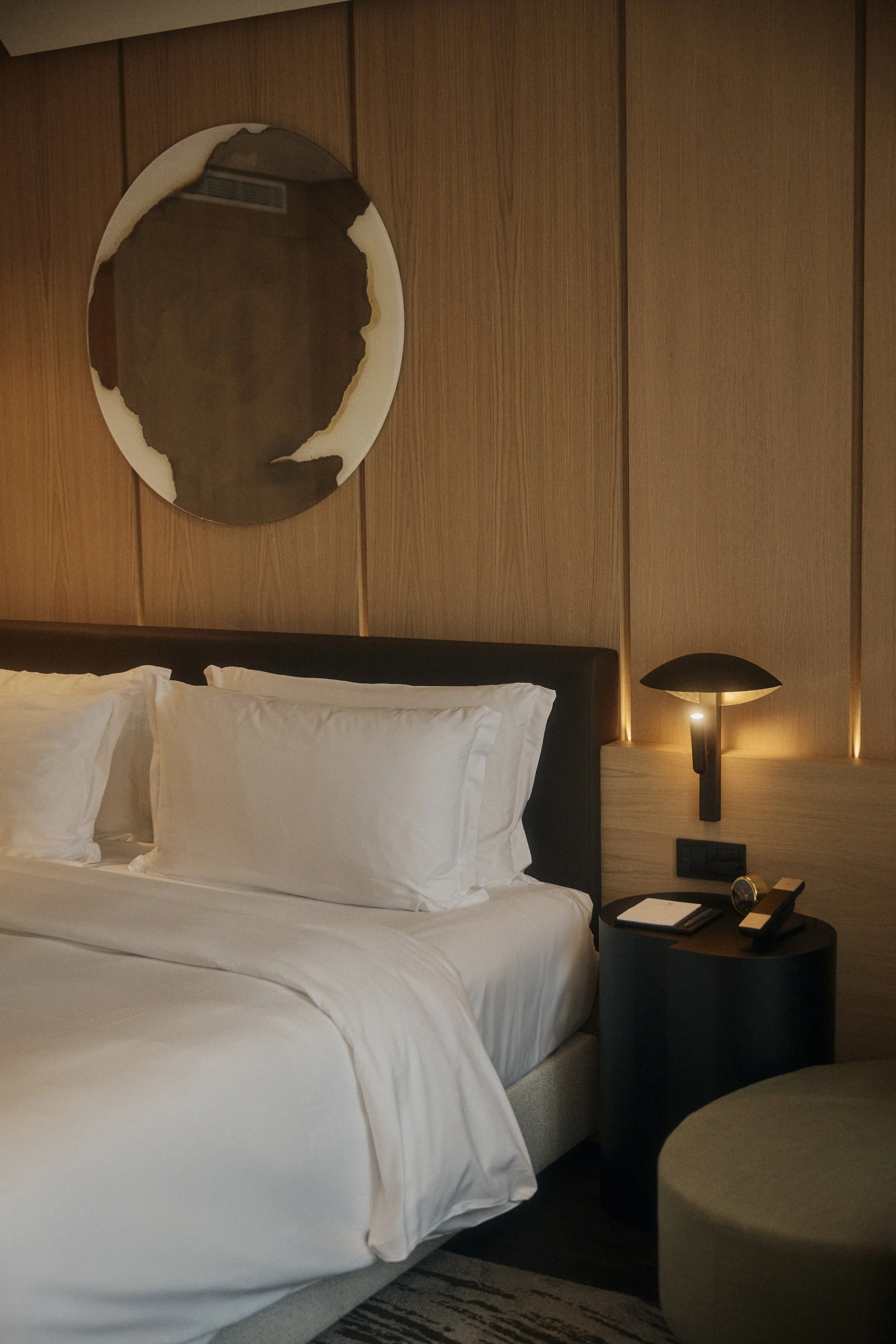 Modernes Hotelzimmer mit Holzwänden, einem Bett mit weißen Kissen, einem runden Spiegel an der Wand, einem Nachttisch mit Lampe und Notizbuch.
