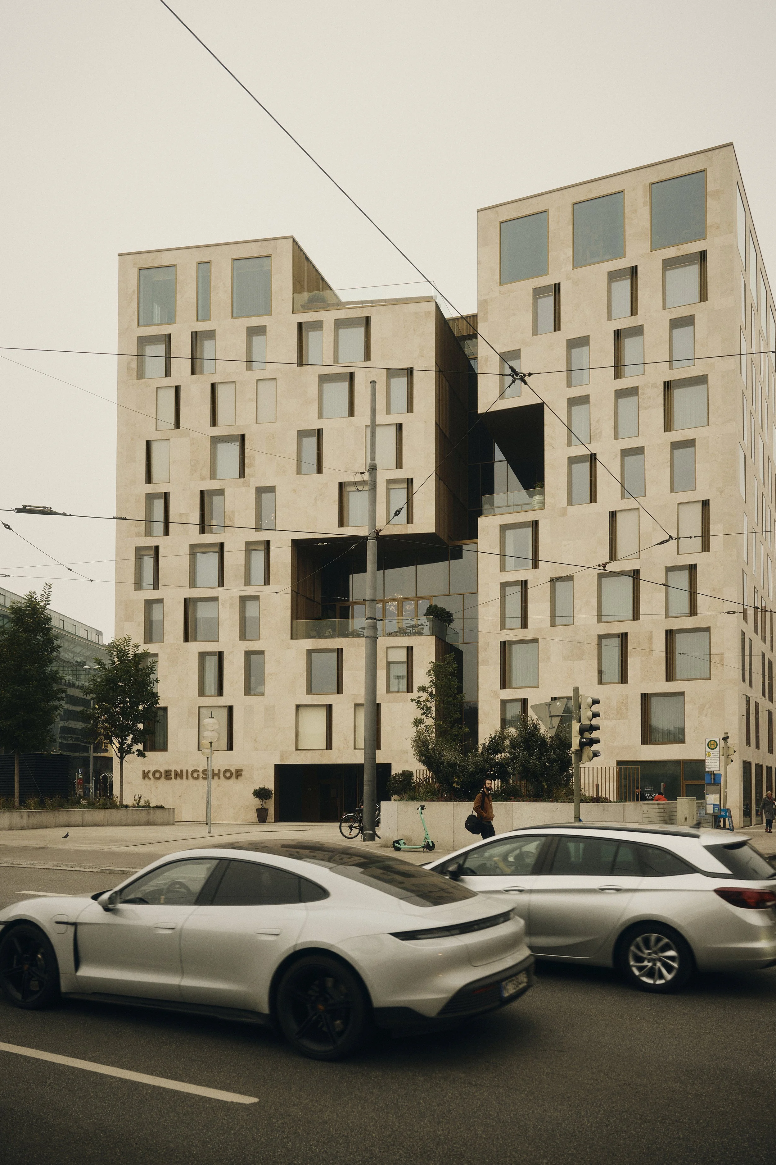 Modernes Gebäude mit geometrischen Formen in urbaner Umgebung, darunter vorbeifahrende Autos, Bäume und Gehweg.