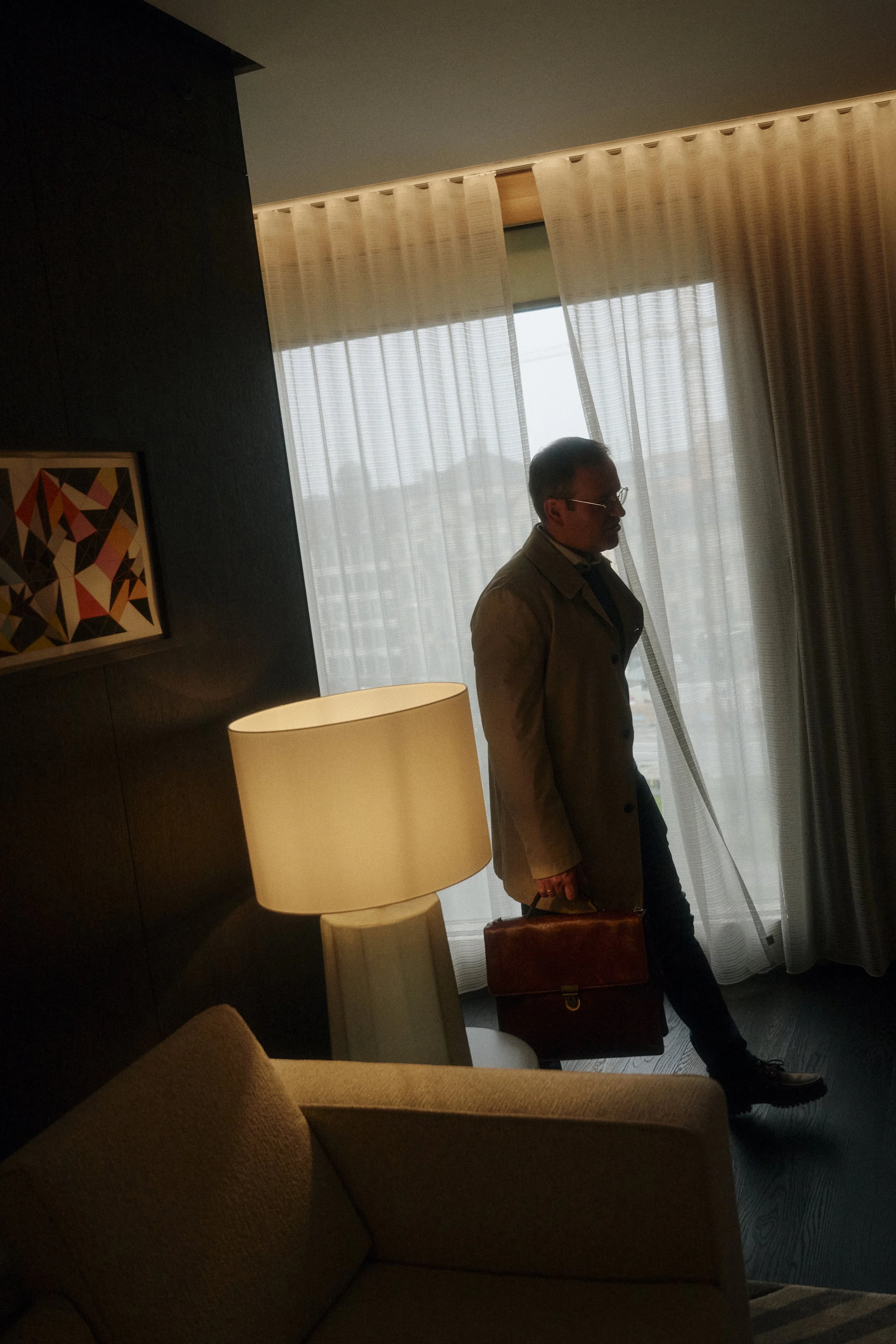Mann in einem Raum mit Vorhängen, Stehlampe und abstraktem Gemälde; hält eine Aktentasche.