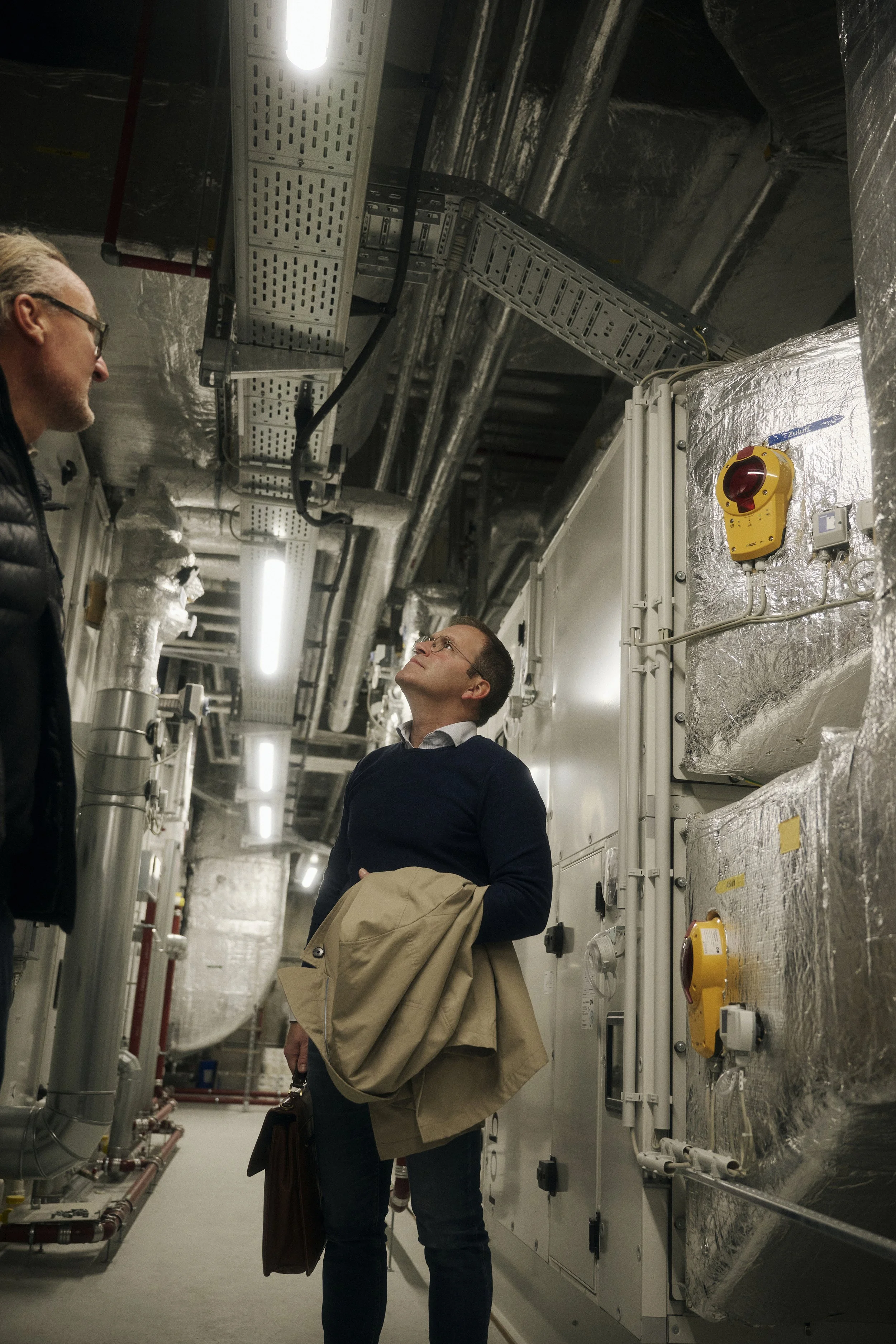 Zwei Personen in einem technischen Raum mit Rohren, Leitungen und Isolierungen an den Wänden und der Decke. Eine Person trägt eine Jacke und hält eine Aktentasche.