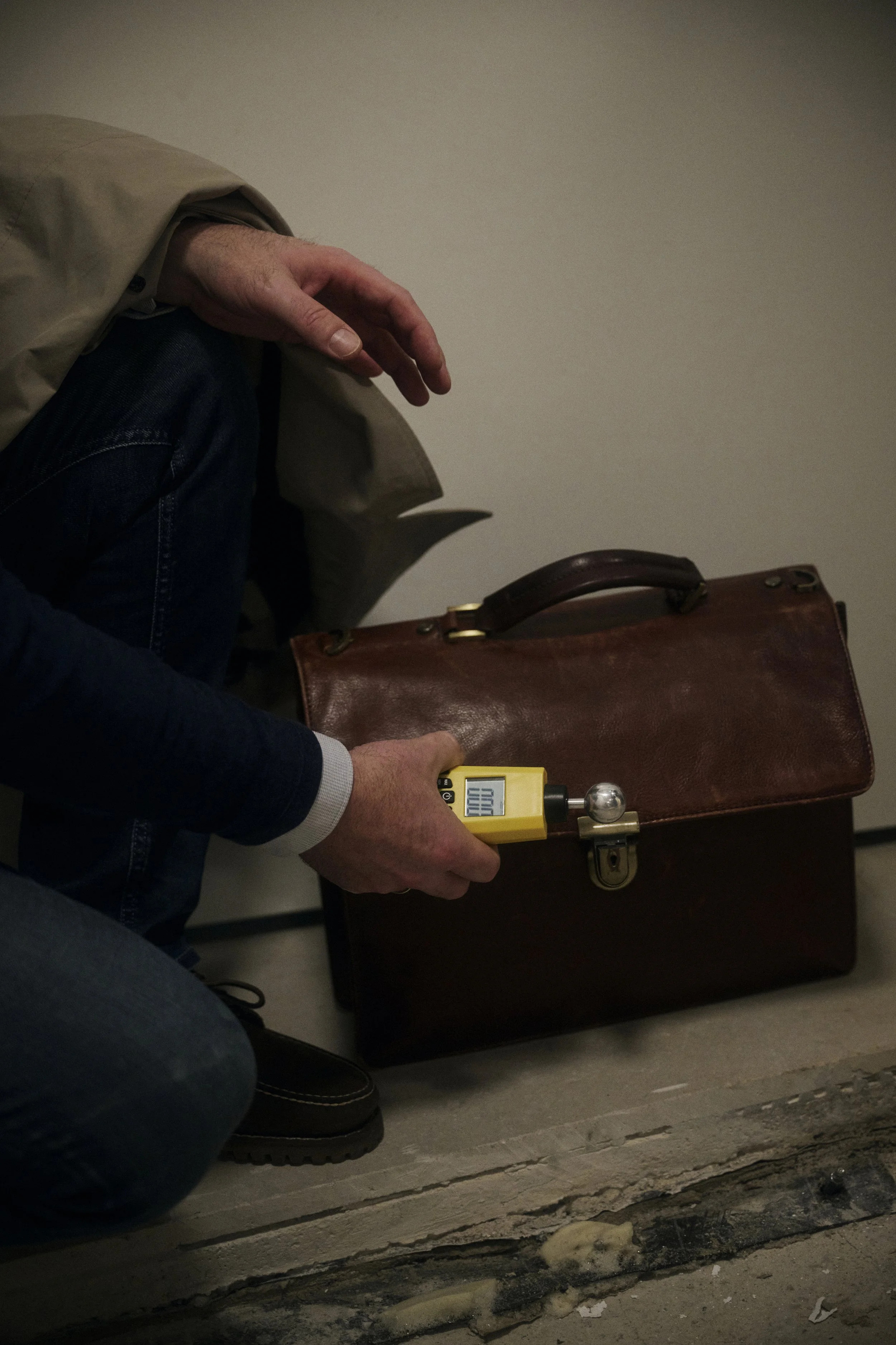 Eine Person in einem Mantel kniet vor einem braunen Lederkoffer und hält ein gelbes Gerät mit Display in der Hand.