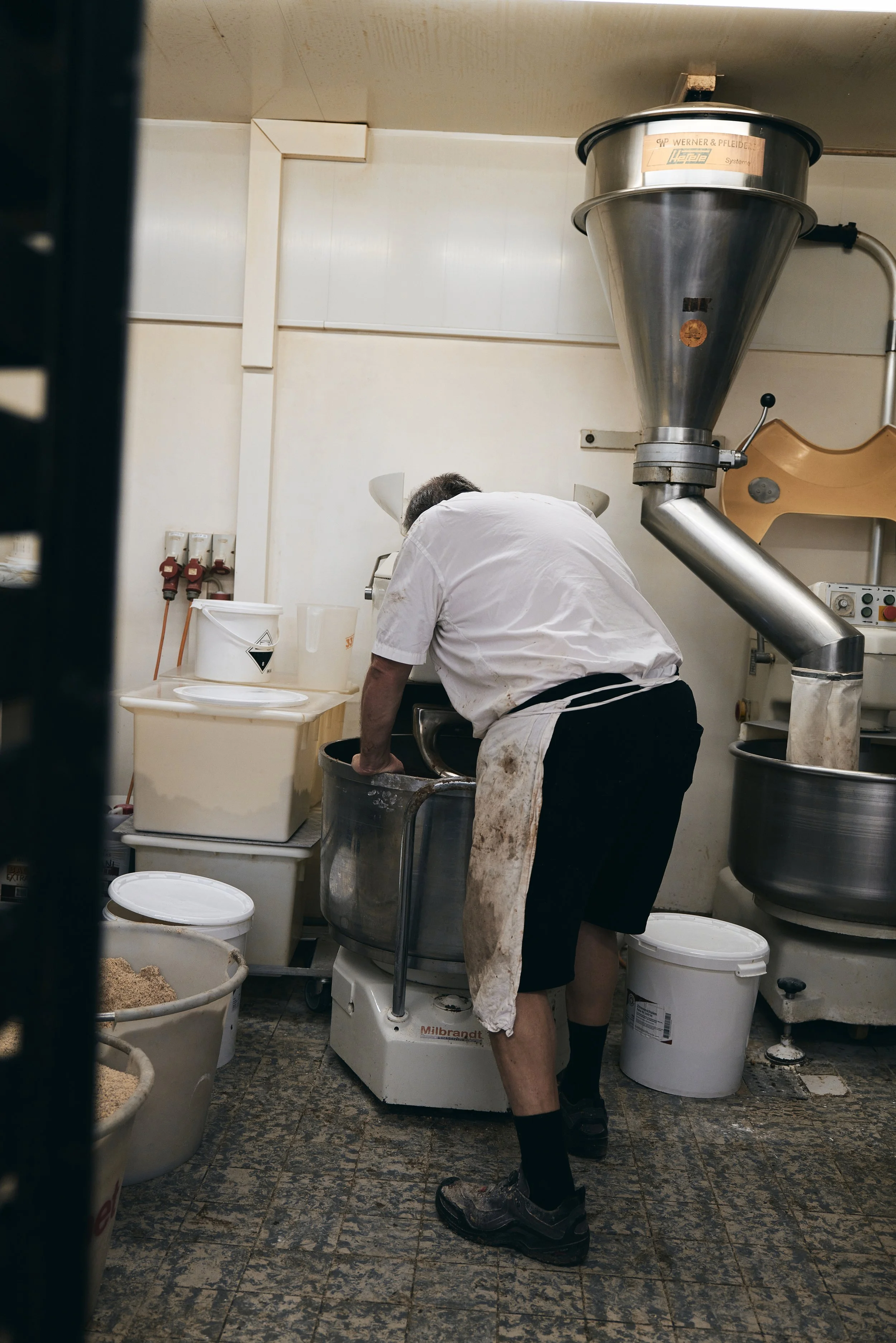 Bäcker bei der Arbeit in einer Bäckerei mit Mischmaschine und Teigbehältern.
