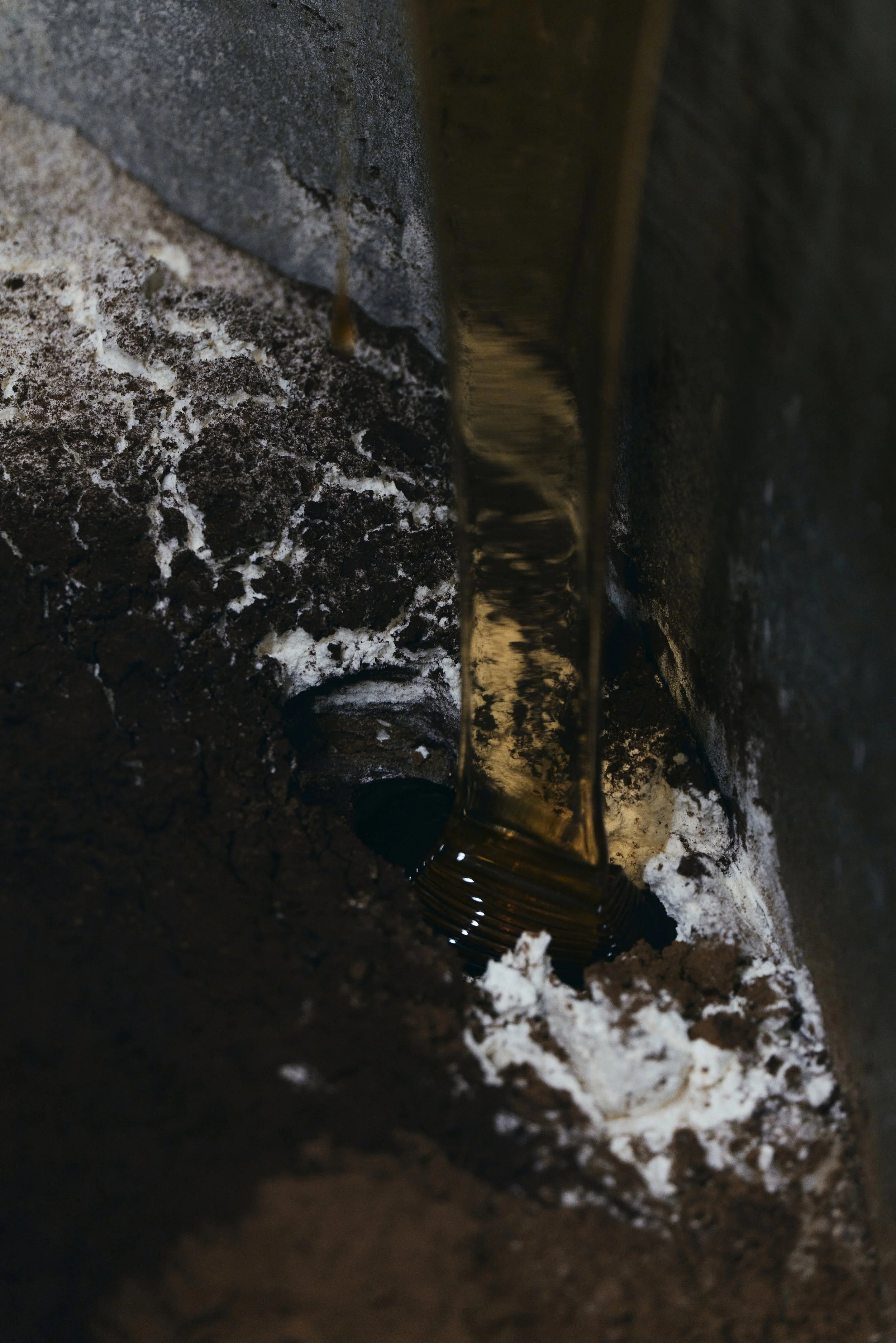 Farbiger Flüssigkeitsstrahl, der auf eine dunkle, strukturierte Oberfläche trifft, umgeben von Trockenmasse oder Staub, möglicherweise Erde oder Kakaopulver.