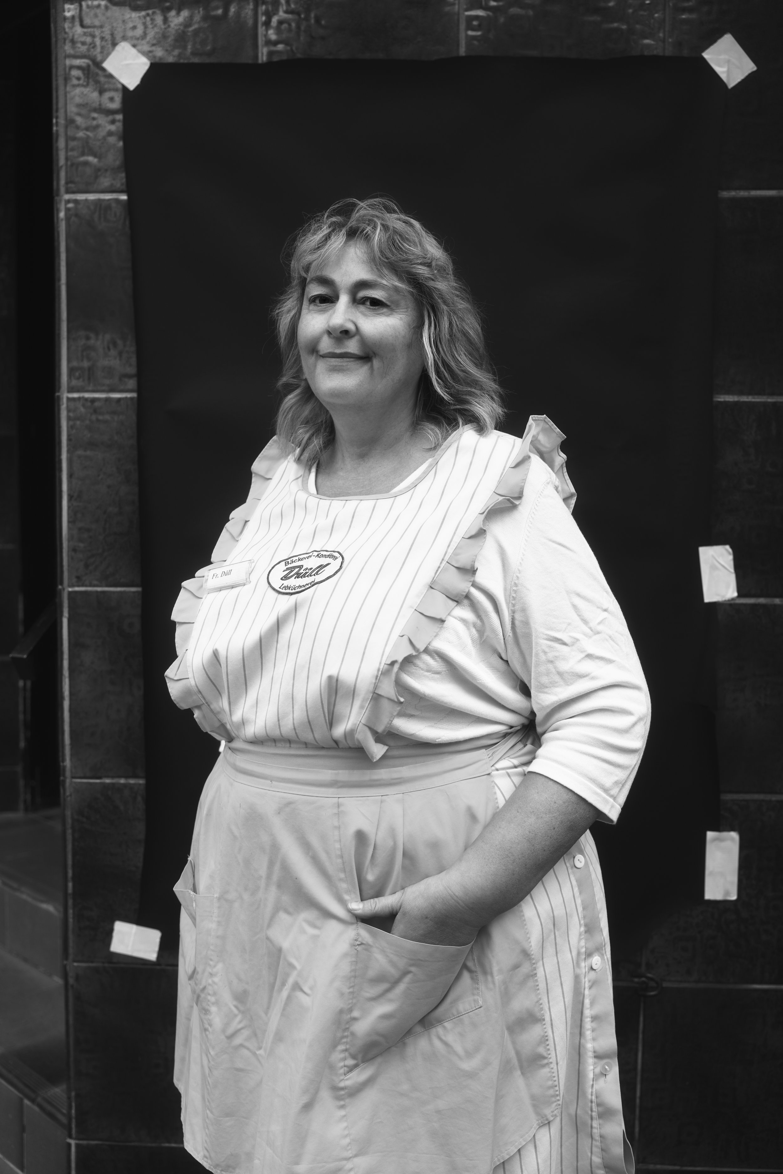 Schwarz-Weiß-Foto einer Frau in einer gestreiften Schürze mit lächelndem Ausdruck, stehend vor einem dunklen Hintergrund.