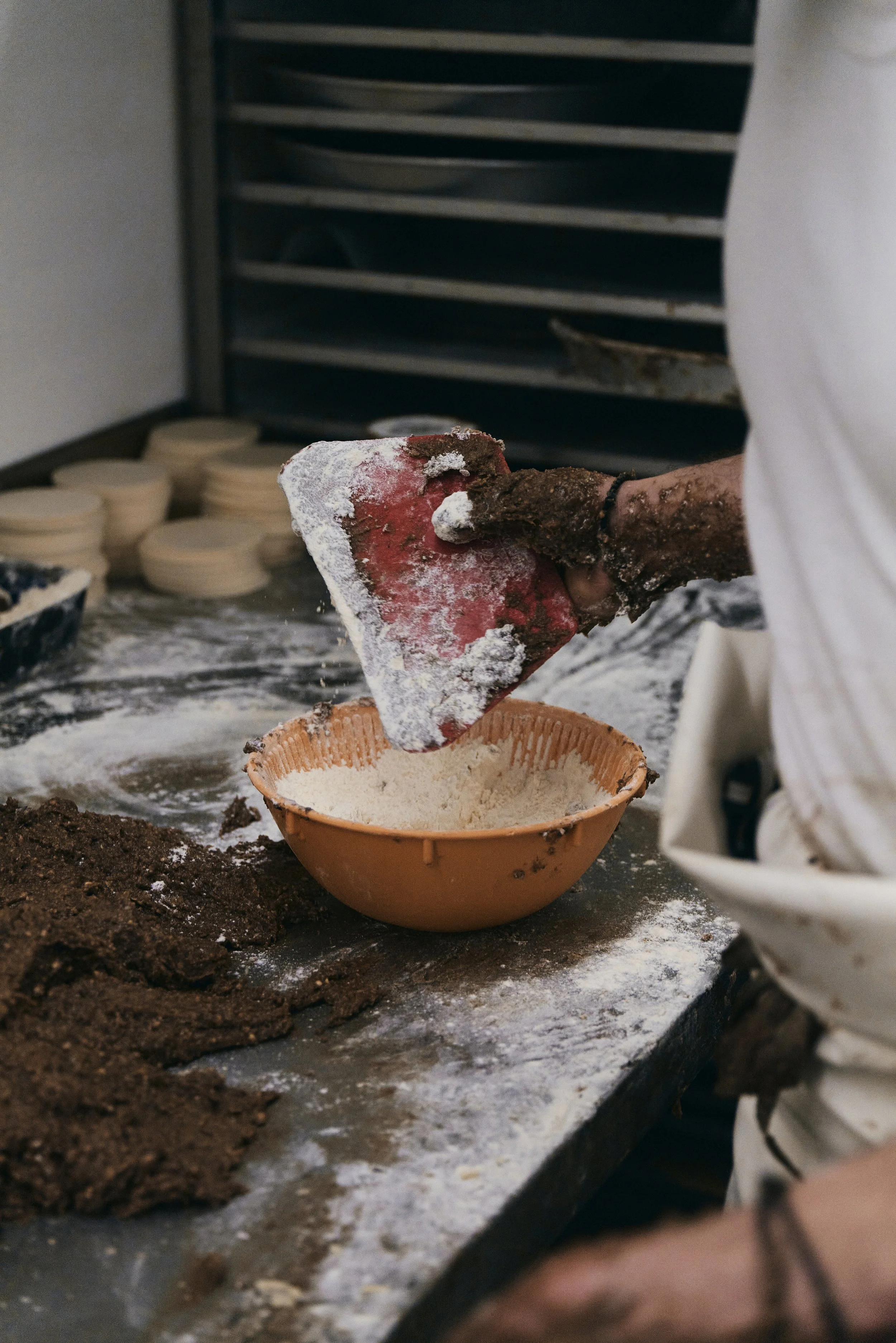 Person beim Backen in einer Bäckerei, hält einen Teigschaber über eine Schüssel mit Mehl, im Hintergrund Teiglinge und Backbleche.