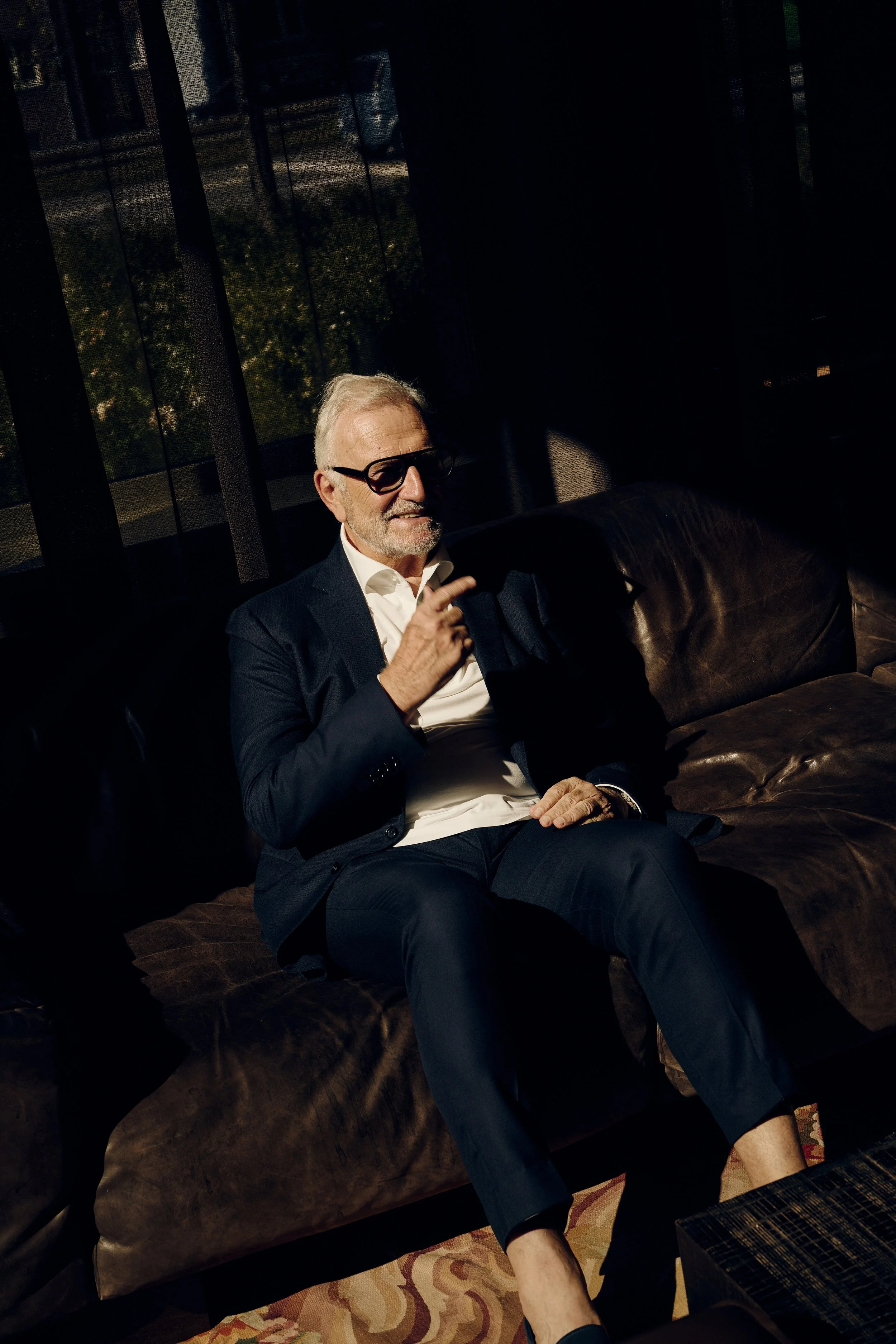 Dieter Müller in Anzug sitzt auf braunem Ledersofa, trägt Sonnenbrille, Finger am Kinn, Licht- und Schattenspiel im Raum.