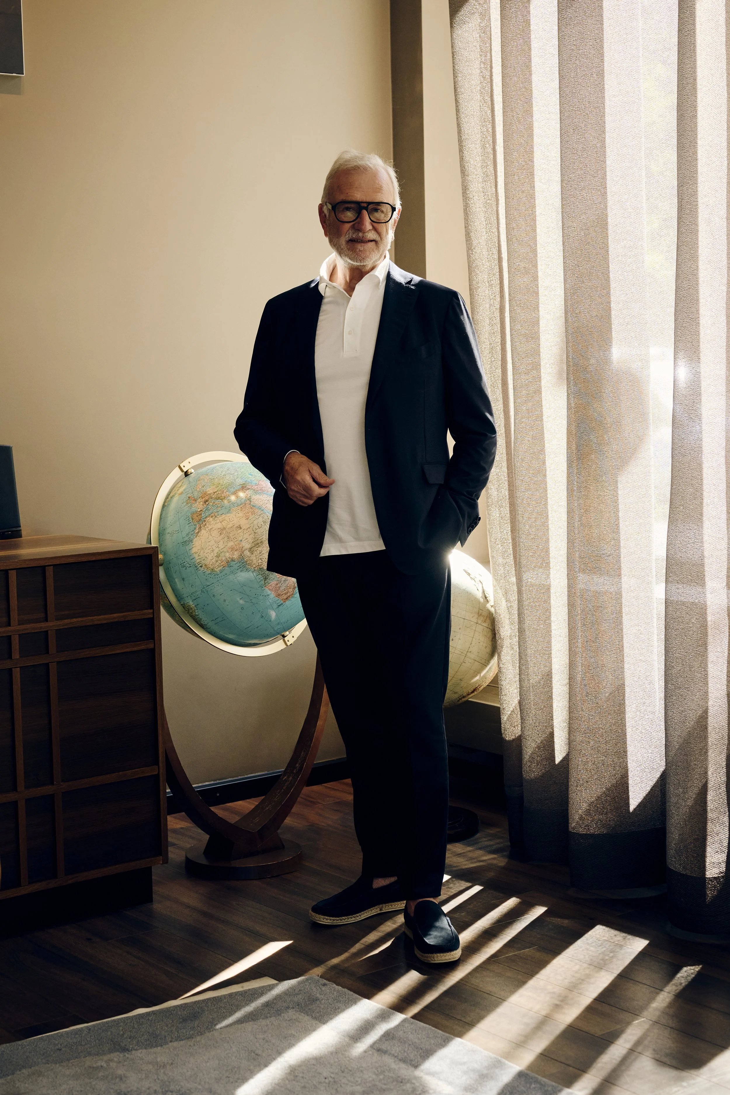 Dieter Müller in einem Anzug steht neben einem Globus in einem Raum mit Holzfußboden und Vorhängen.