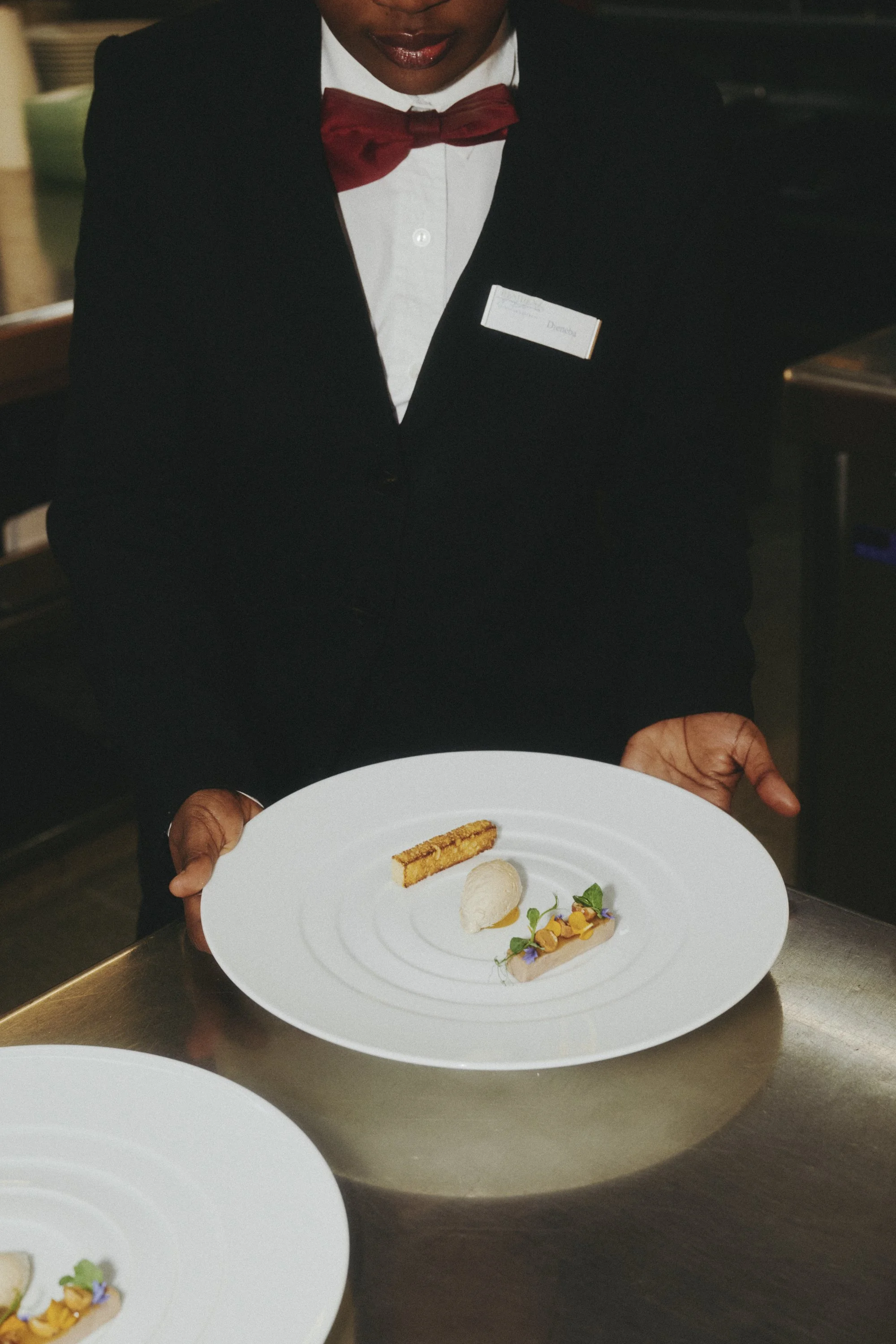 Ein Kellner in schwarzem Anzug und roter Fliege hält einen Teller mit einer eleganten Dessertpräsentation, bestehend aus einer kleinen Eiskugel, einem Stück Kuchen und dekorativen Blüten und Kräutern.