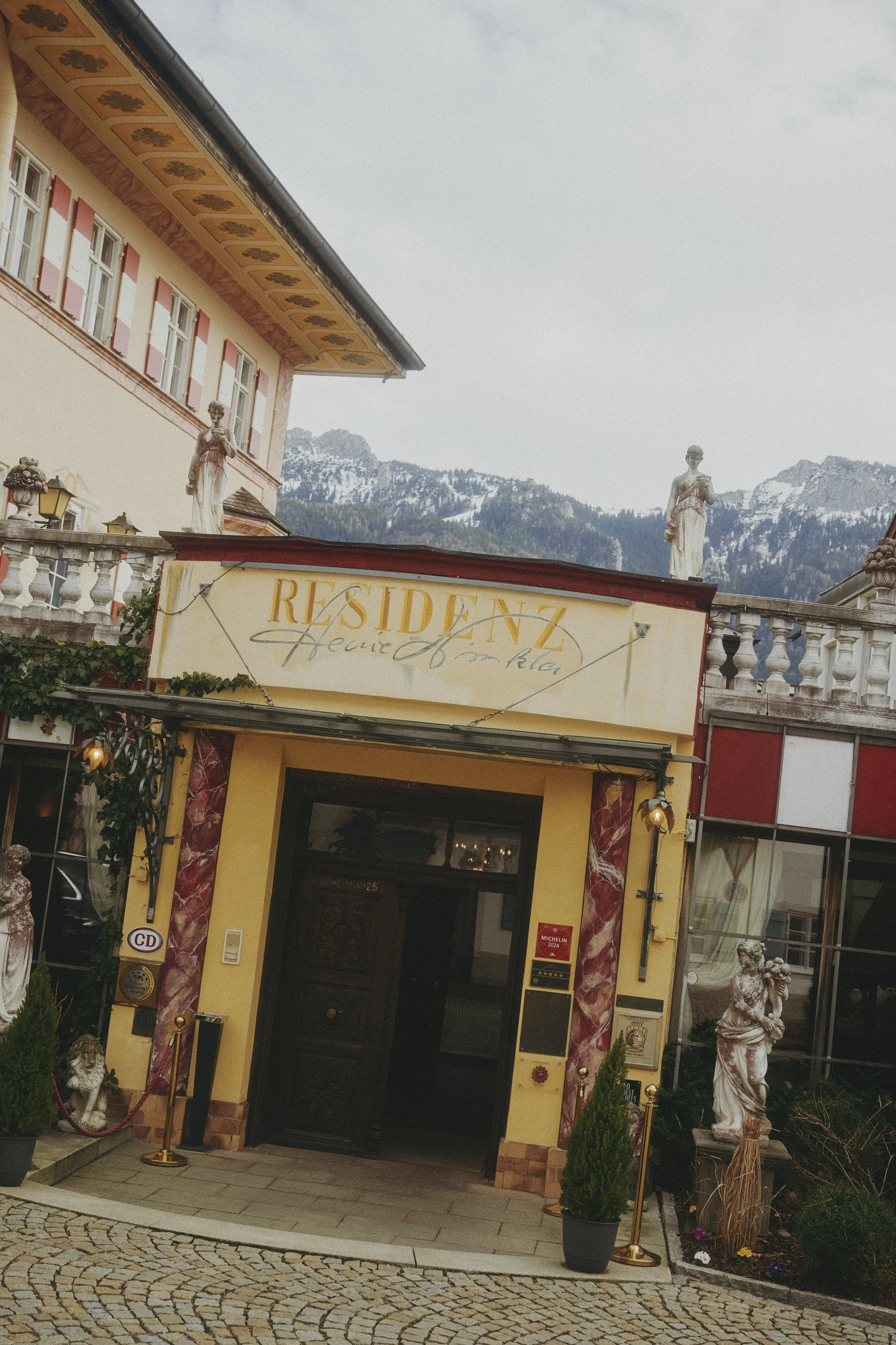 Eingang eines eleganten Gebäudes mit der Aufschrift "Residenz" über der Tür, flankiert von Statuen und Topfpflanzen. Im Hintergrund sind schneebedeckte Berge sichtbar.