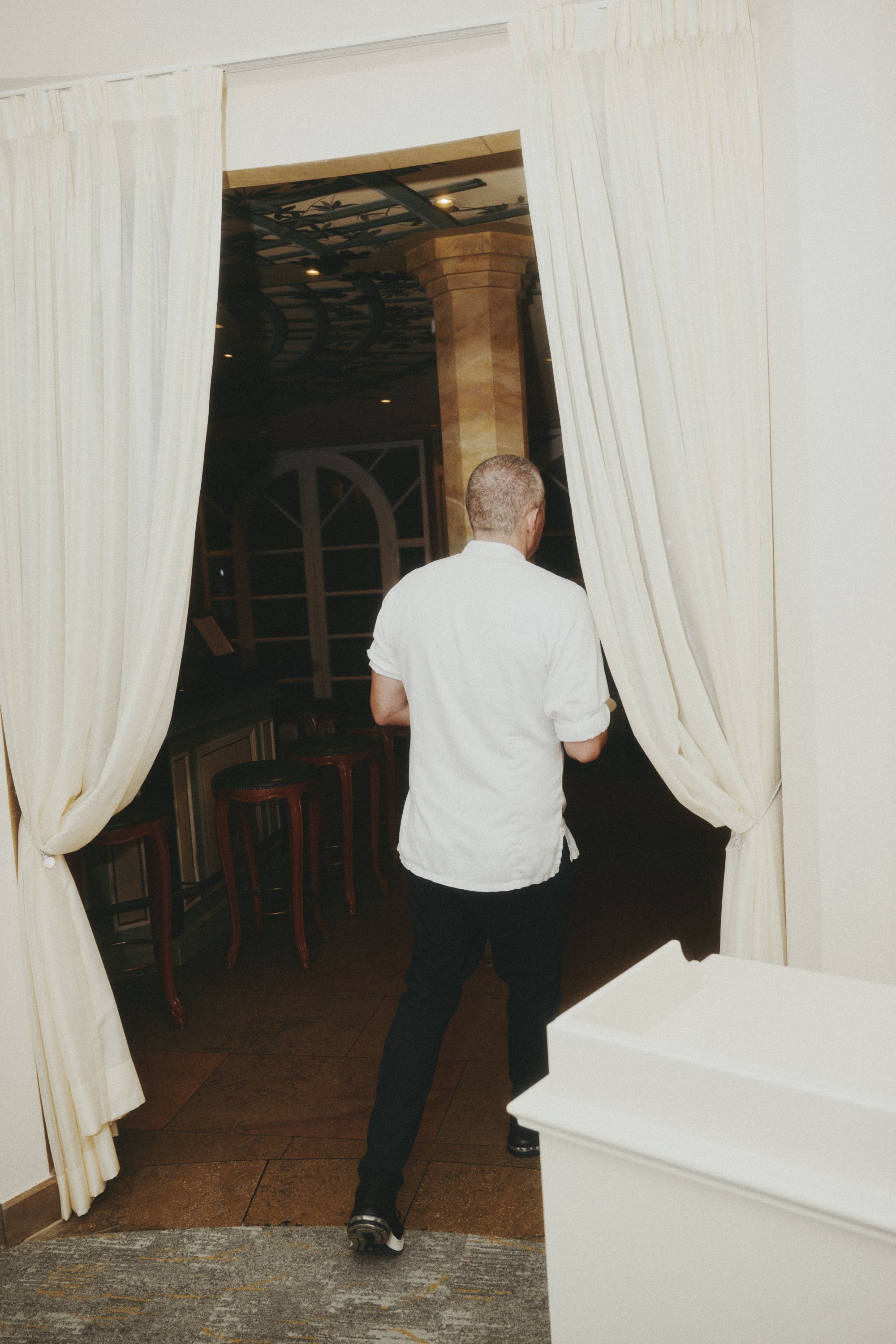 Rückansicht einer Person in weißem Hemd, die durch einen Vorhang in einen dunklen Raum mit Barhockern geht.