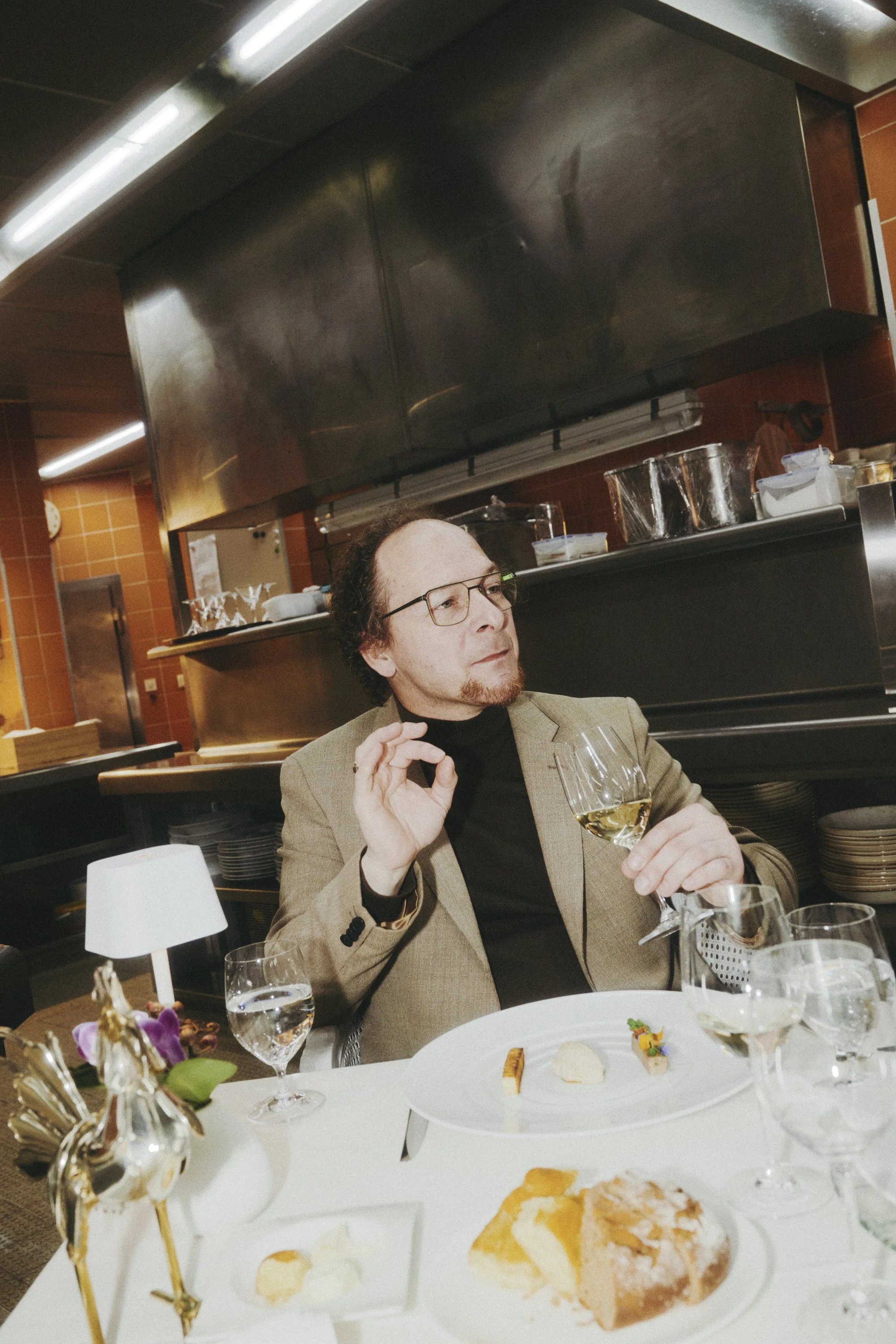 Ein Mann in einem Restaurant hält ein Glas Weißwein und sitzt an einem Tisch mit verschiedenen Speisen. Im Hintergrund ist eine professionelle Küchenumgebung zu sehen. Der Mann trägt einen hellen Anzug und eine Brille.