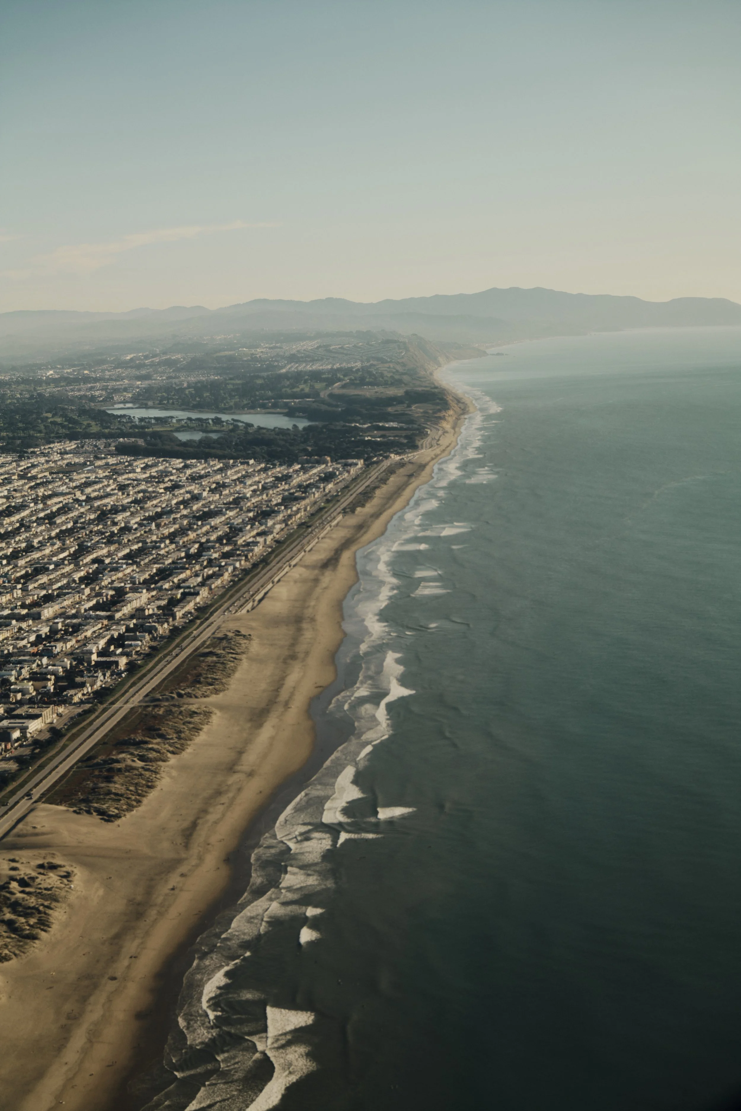 Luftaufnahme eines langen Sandstrandes mit anliegenden Stadtgebieten und Hügeln im Hintergrund, Meer rechts im Bild.