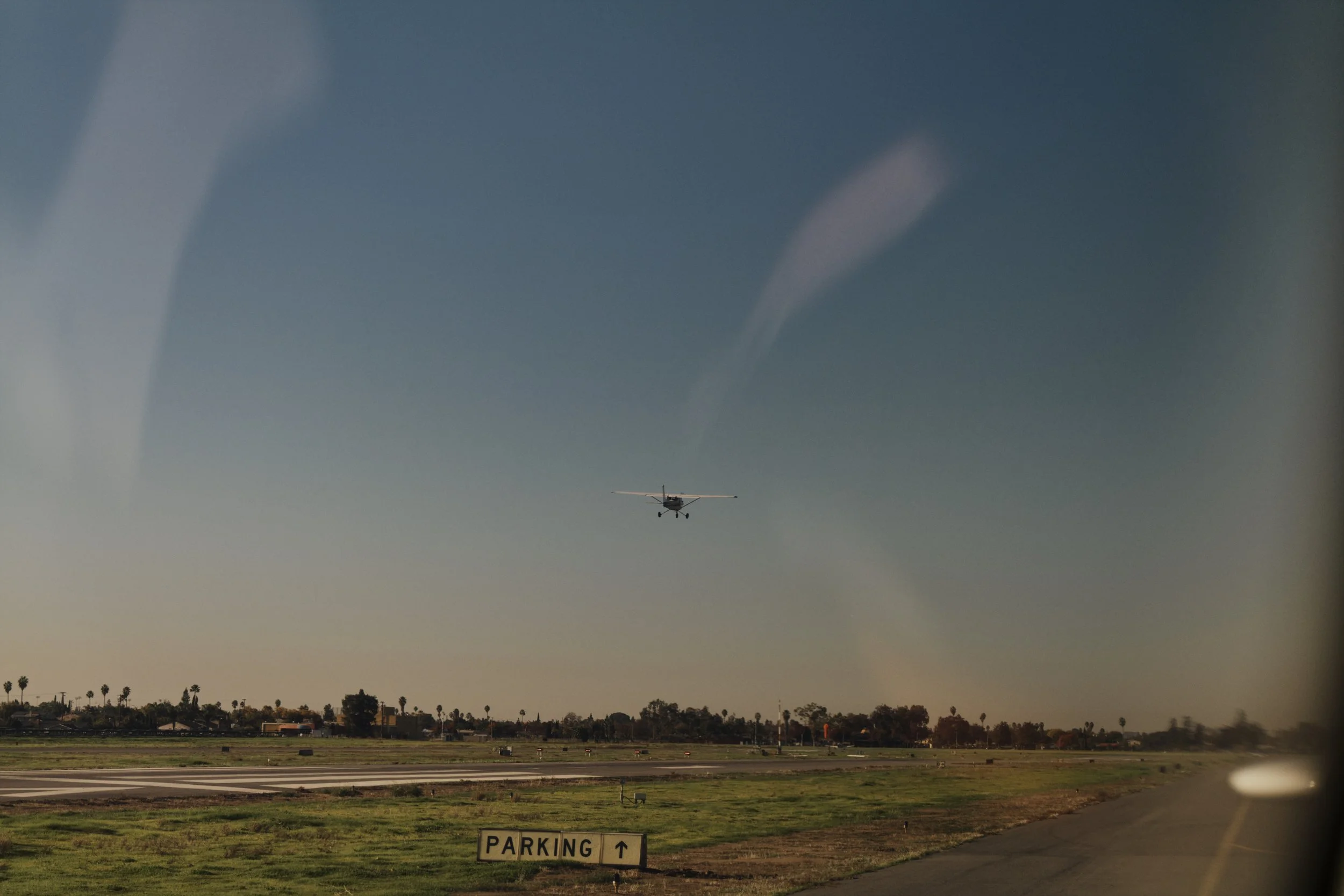 Kleines Flugzeug im Landeanflug auf eine Landebahn mit Parkhinweisschild, umgeben von Feldern und Bäumen, blauer Himmel im Hintergrund.