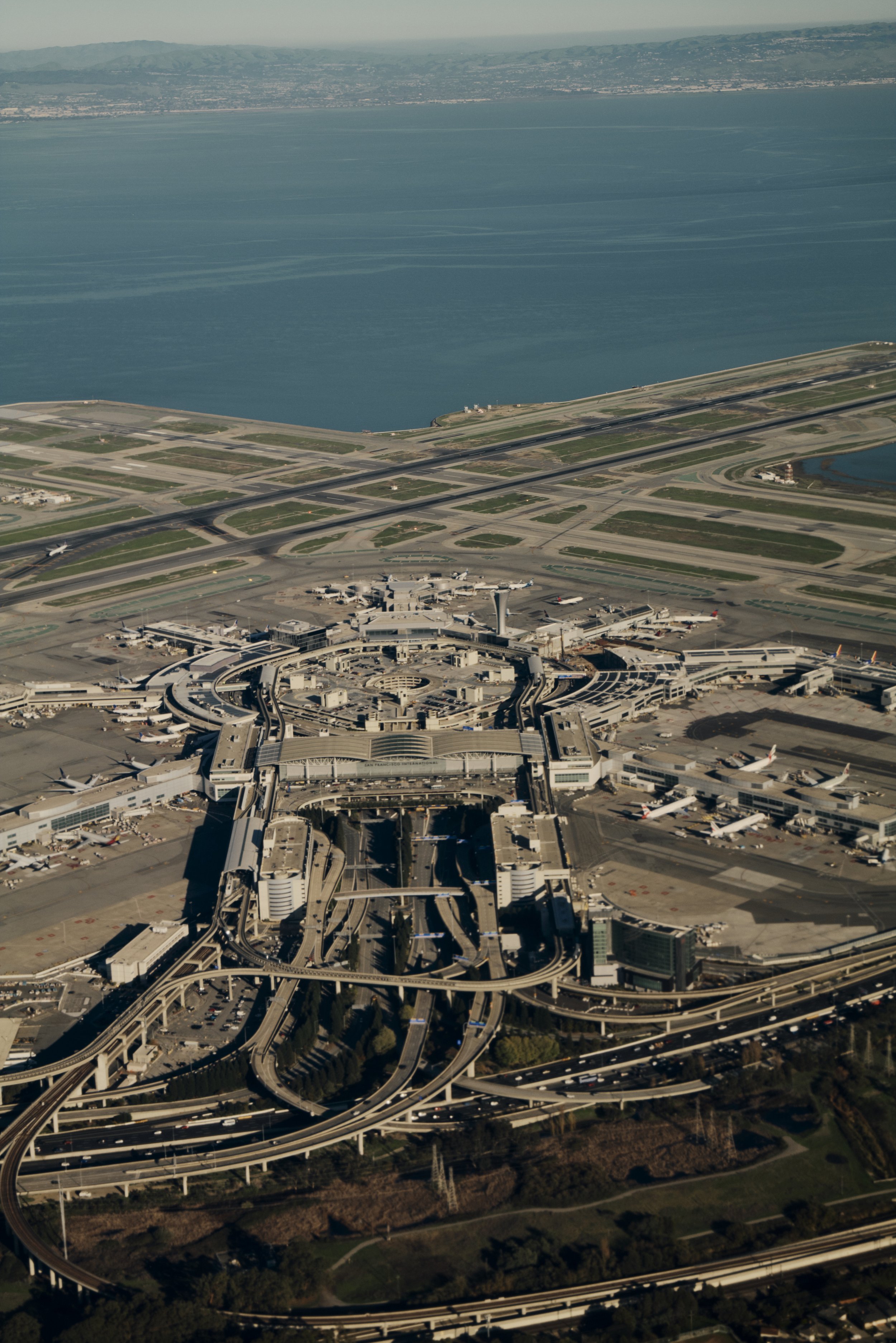 Luftaufnahme eines internationalen Flughafens mit mehreren Rollbahnen, Flugzeugen auf Rollfeldern, einem zentralen Terminalkomplex und Zufahrtsstraßen im Vordergrund, umgeben von Wasser und Land.