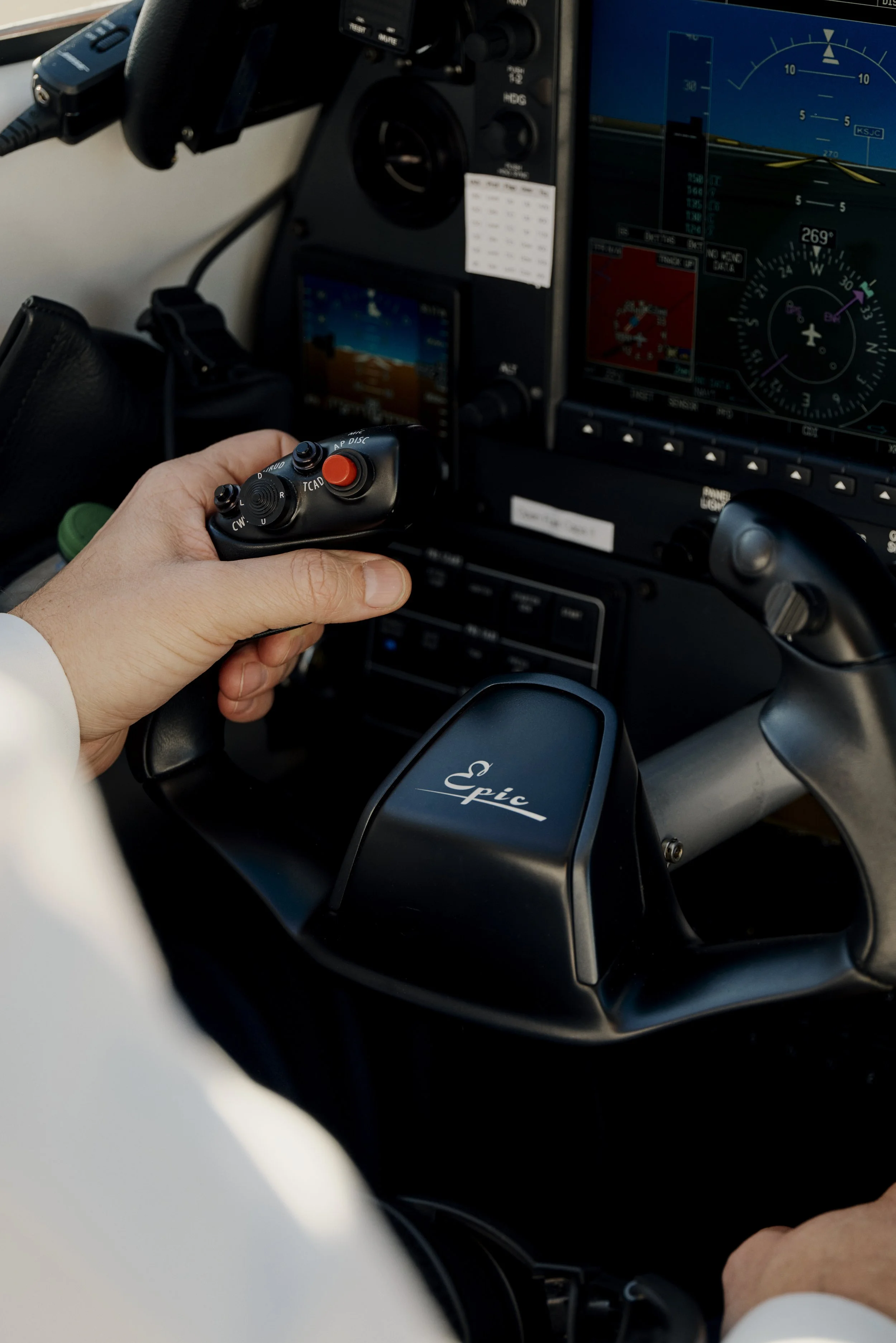 Nahaufnahme eines Cockpit-Steuerhorns mit der Aufschrift "Epic". Eine Hand hält einen Steuerhebel mit mehreren Knöpfen und einem roten Knopf. Im Hintergrund sind Instrumentenanzeigen und ein Navigationsbildschirm zu sehen.