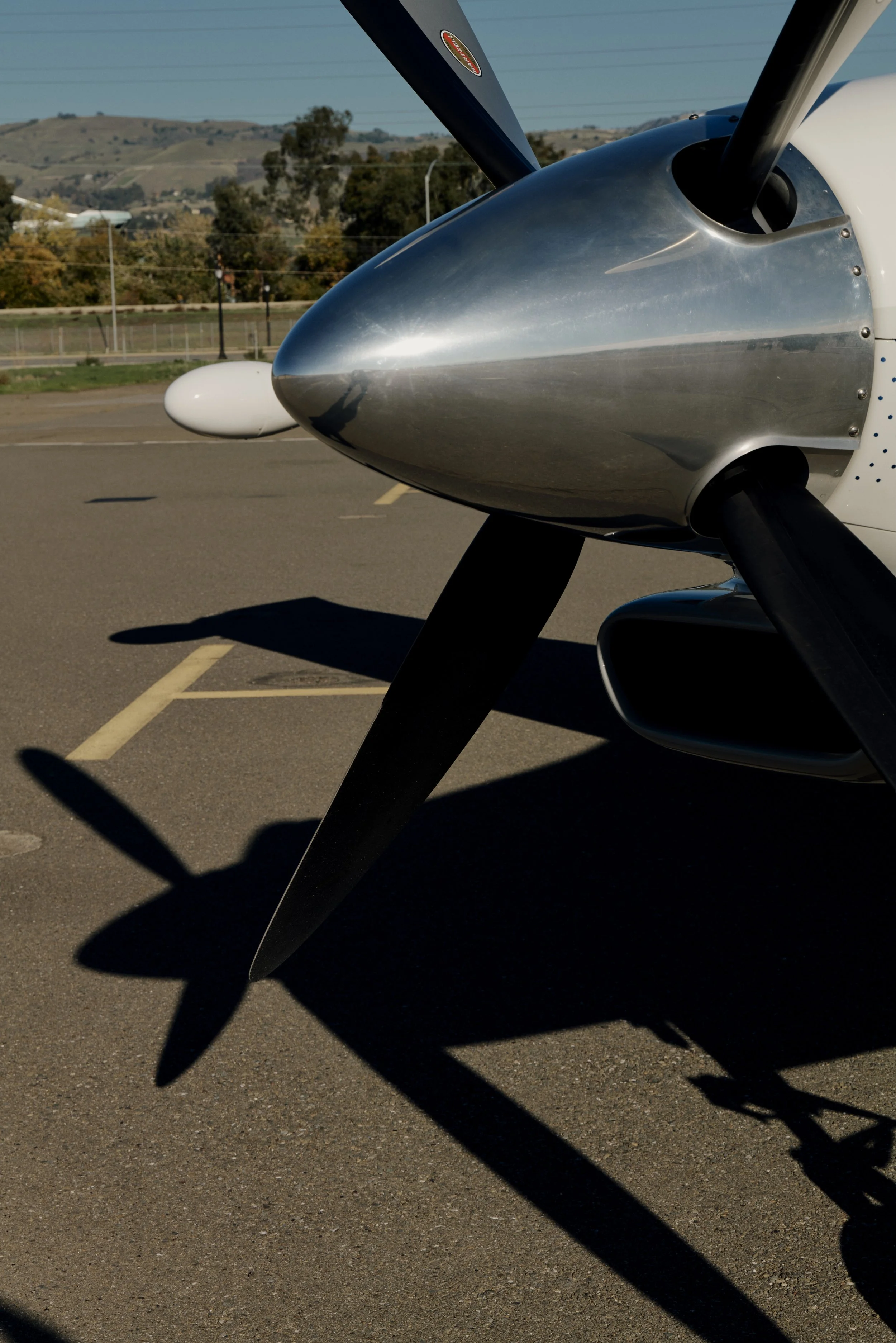 Nahaufnahme eines Propellers eines Flugzeugs auf einem Rollfeld, mit Schatten des Propellers auf dem Boden und Landschaft im Hintergrund.