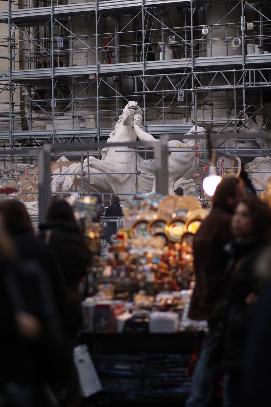 Eine Baustelle mit einem Gerüst, hinter dem eine große, teilweise verhüllte Skulptur eines Pferdes und eines Mannes sichtbar ist. Im Vordergrund unscharf sind Menschen und Souvenirstände.