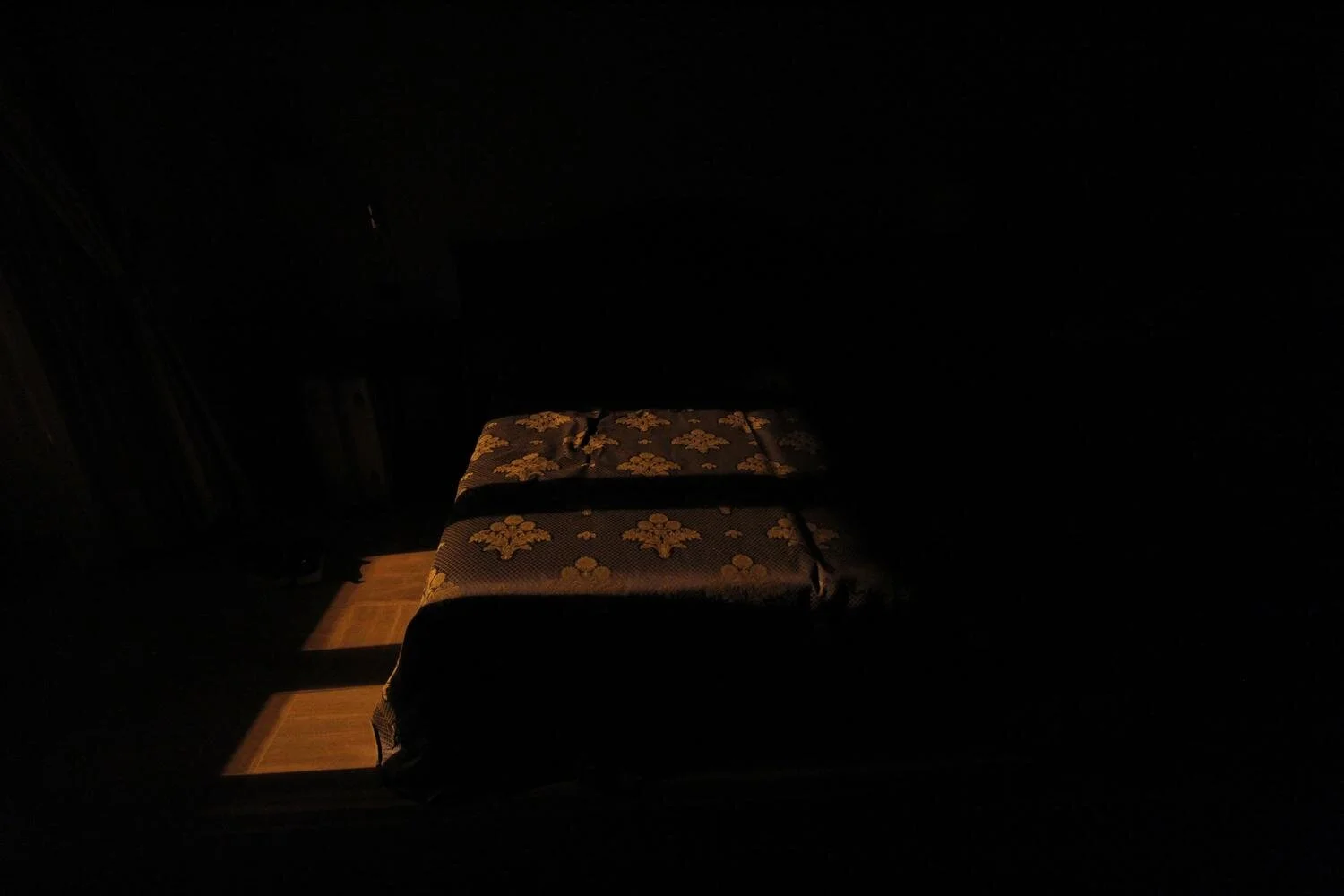 Ein dunkles Zimmer mit einem Bett, auf das etwas Licht fällt. Das Bett hat eine gemusterte Bettdecke.