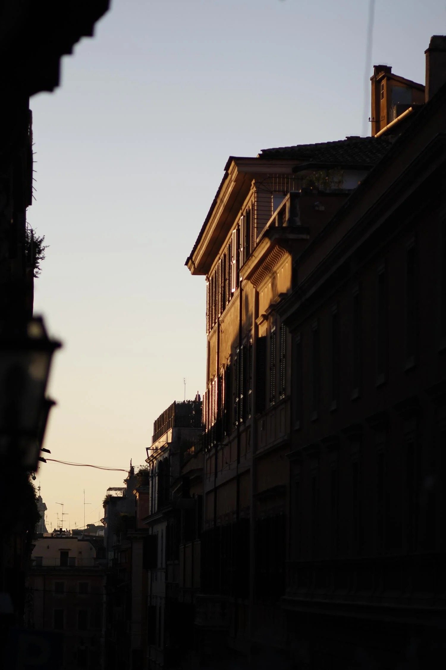 Silhouette einer Stadtstraße im Abendlicht, mit historischem Gebäude und Fensterläden, beleuchtet von der tiefstehenden Sonne.