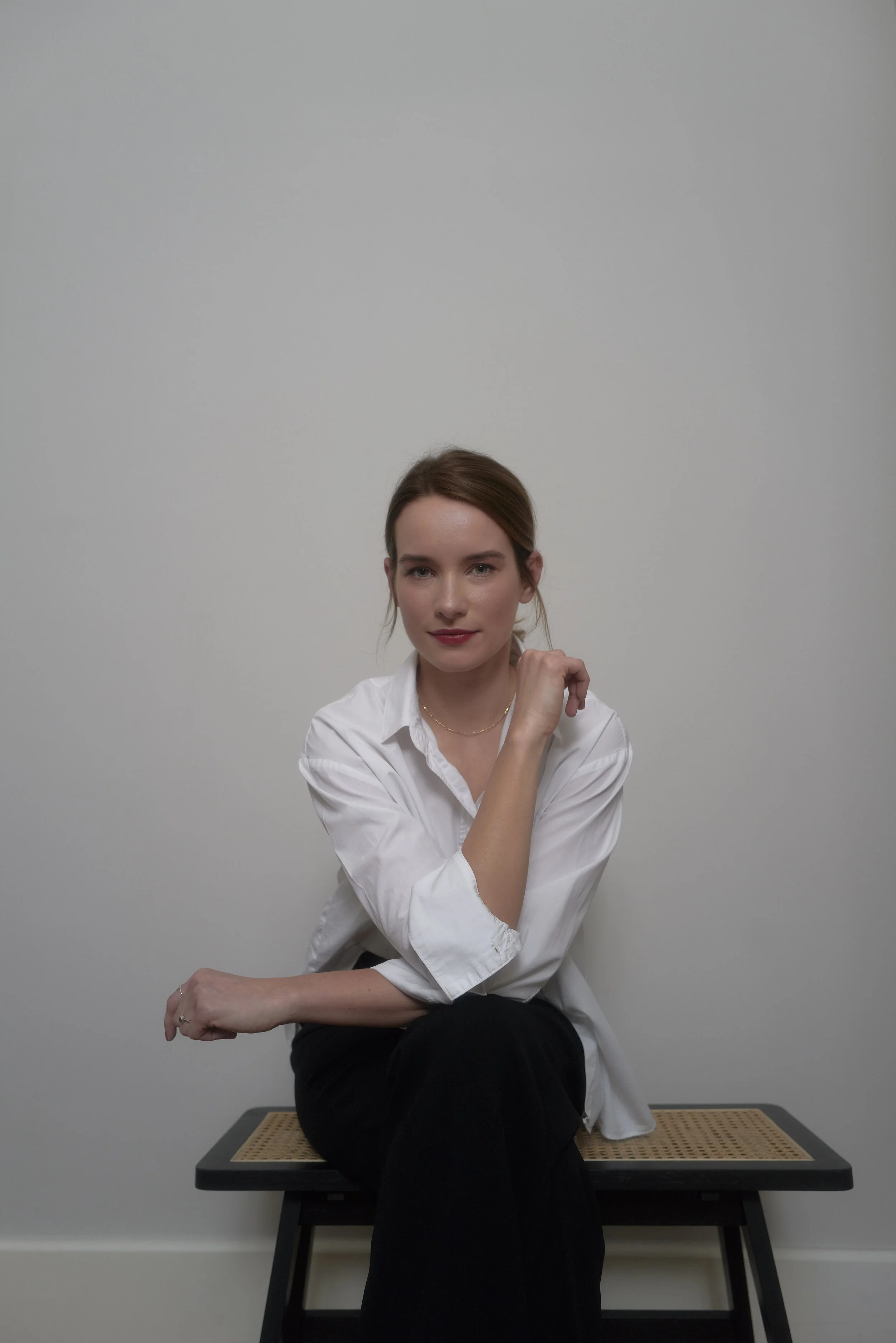 Eine Frau in einem weißen Hemd sitzt auf einem Stuhl vor einem neutralen Hintergrund.