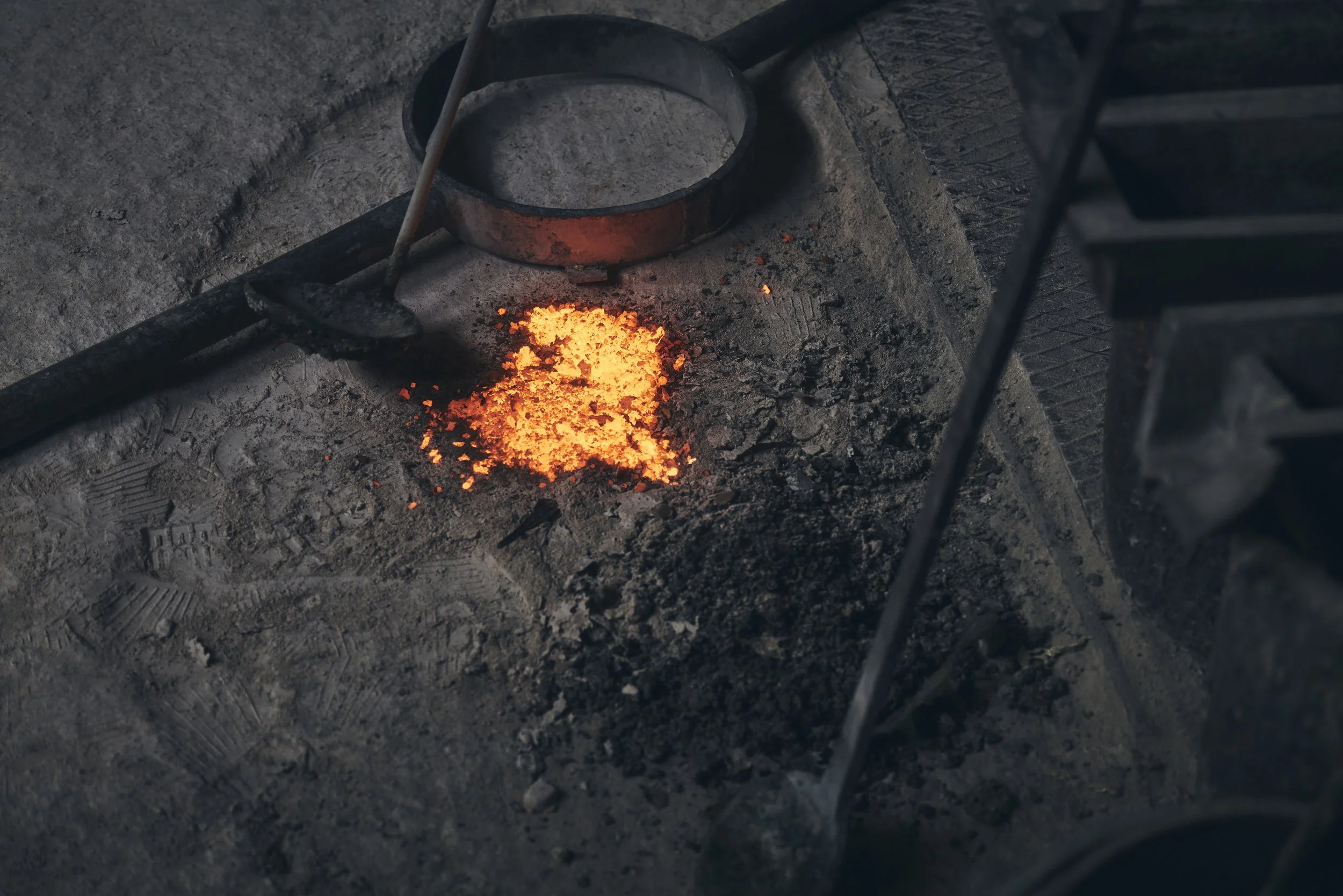 Glühende Kohlen auf einem Betonboden in einer Schmiede mit Werkzeugen, darunter eine Schaufel und ein Metallring.