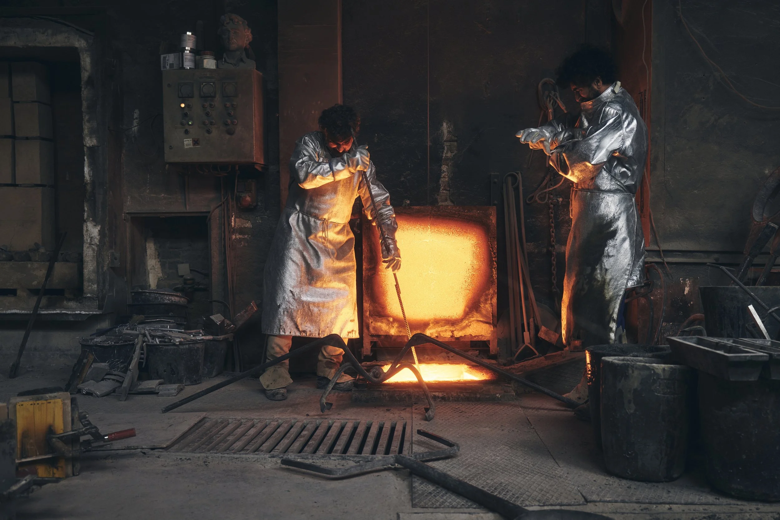 Zwei Arbeiter in Schutzkleidung stehen vor einem Schmelzofen in einer Industrieumgebung. Der Ofen strahlt intensives Licht aus, und die Umgebung ist mit Werkzeugen und Geräten ausgestattet.