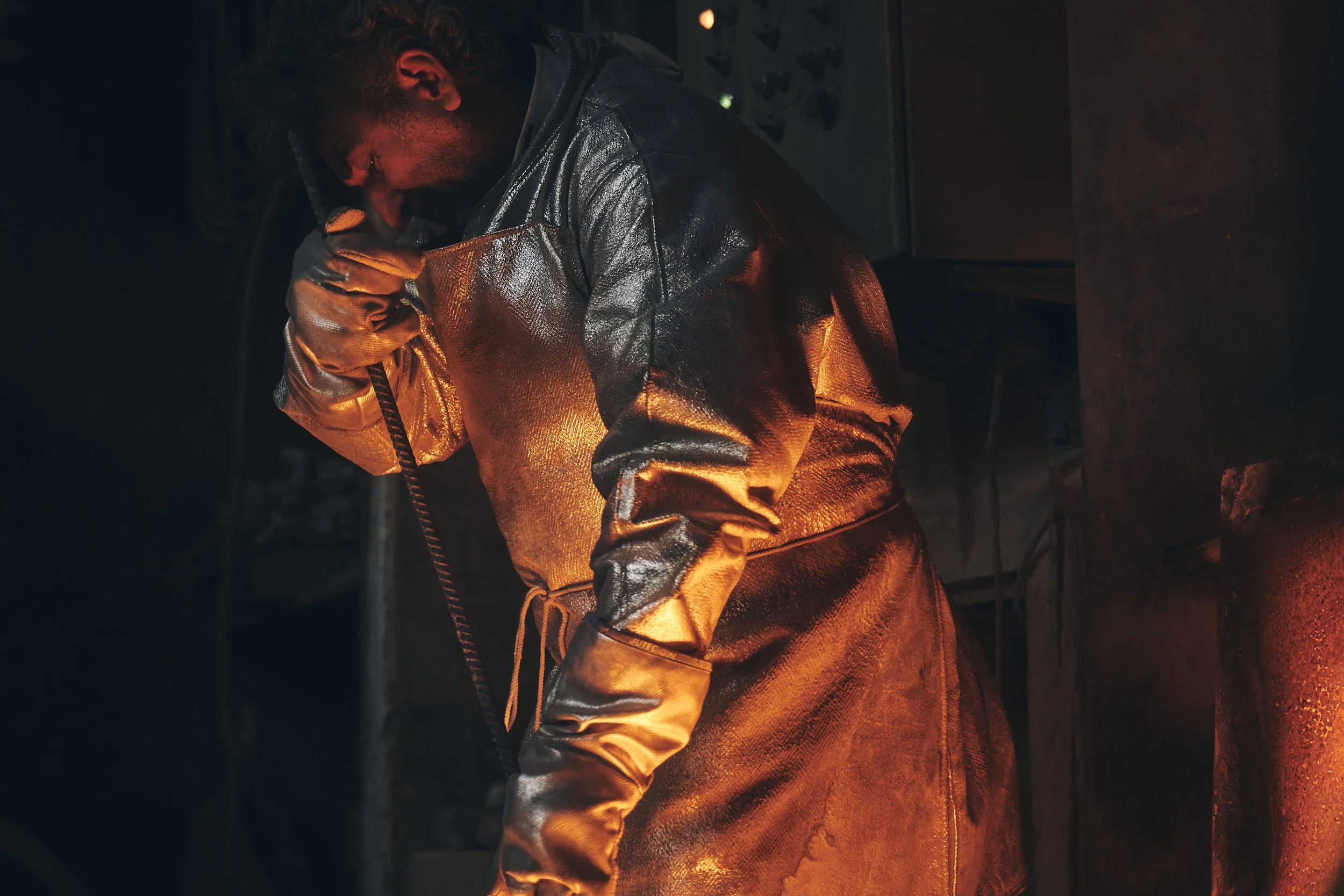 Ein Arbeiter in feuerfester Schutzkleidung in einer dunklen industriellen Umgebung. Die Szene wird von orangefarbenem Licht beleuchtet, das vermutlich von einem glühenden Metall ausgeht.