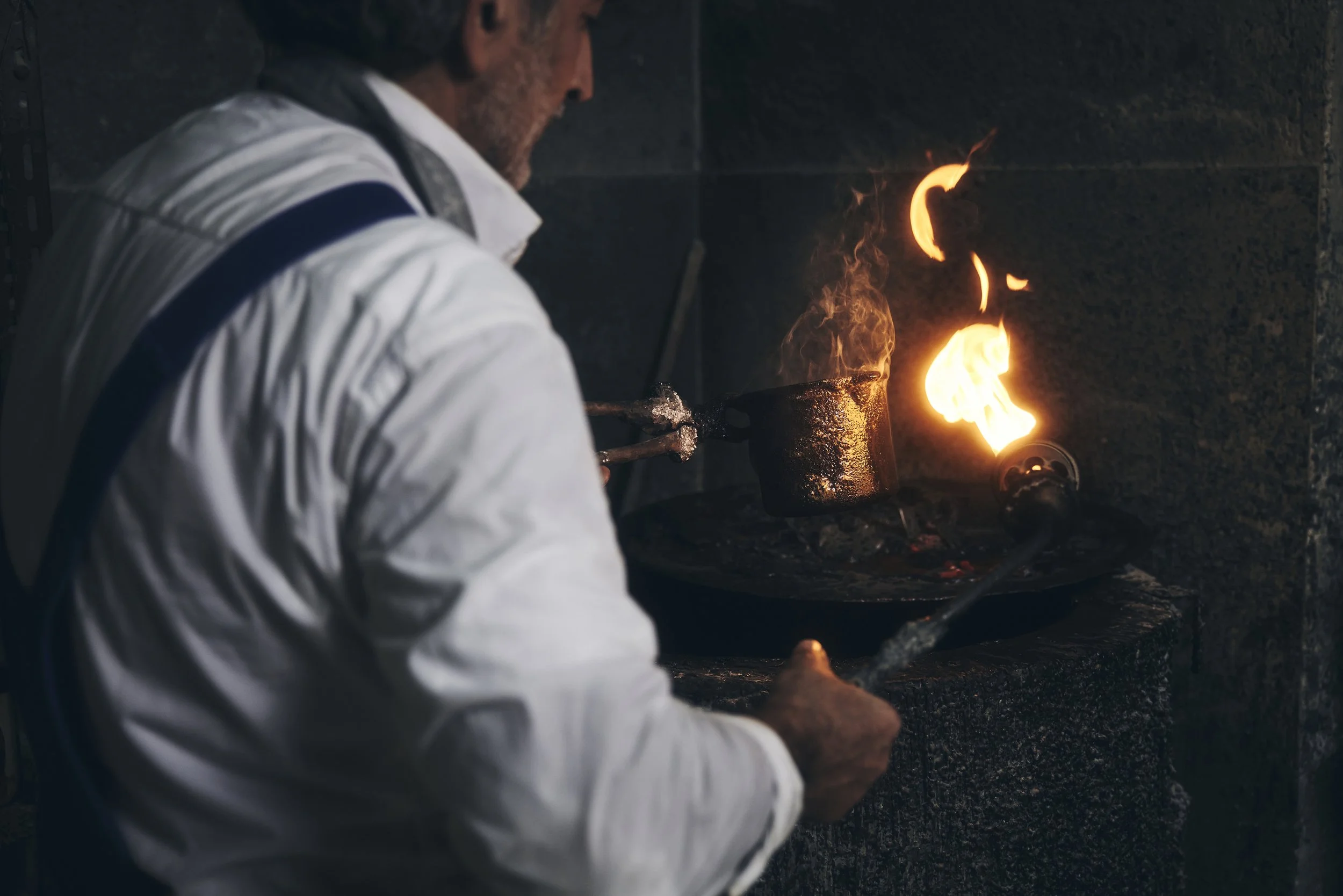 Eine Person in weißem Hemd arbeitet mit einer glühenden Metallmischung über einer offenen Flamme.