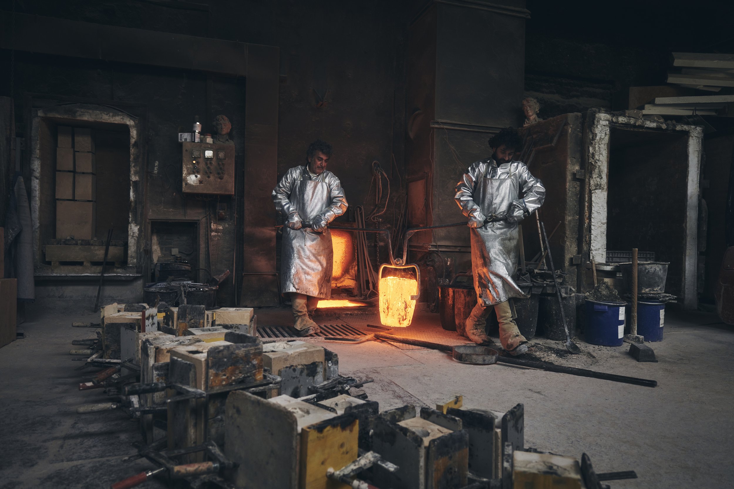 Zwei Arbeiter in Hitzeschutzanzügen in einer Gießerei mit glühendem Metall aus einem Ofen.