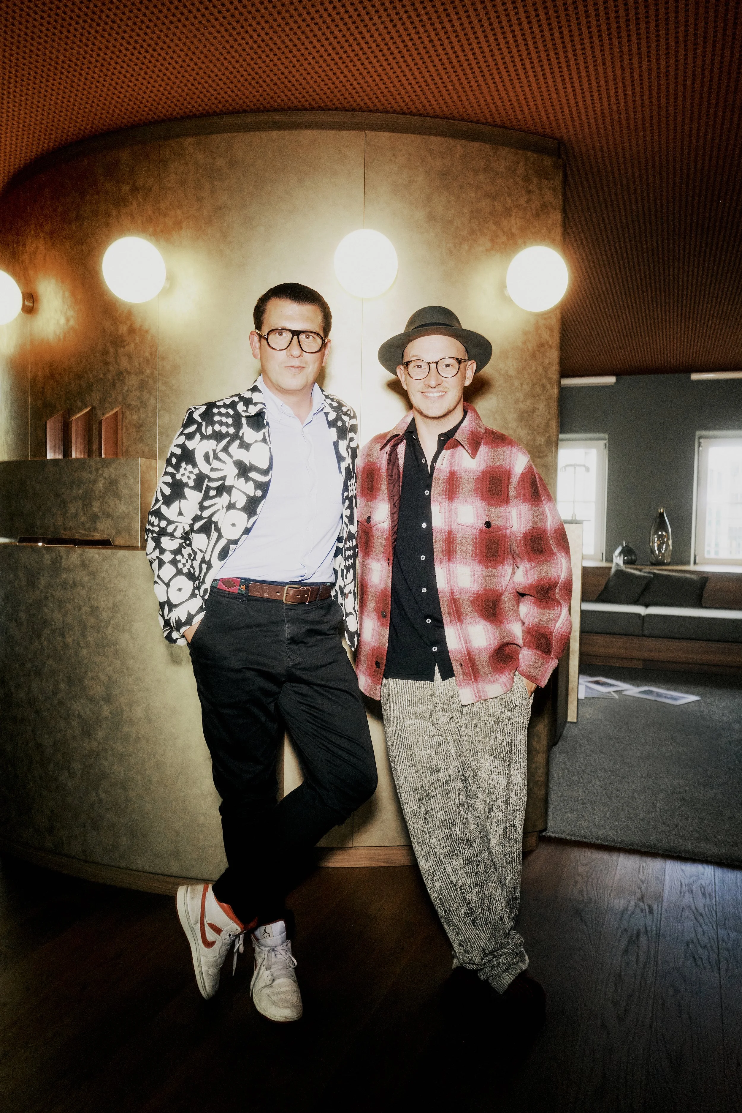 Zwei Männer in modischer Kleidung stehen in einem stilvollen Innenraum, beleuchtet von runden Wandlampen. Einer trägt eine gemusterte Jacke und weiße Sneakers, der andere einen karierten Mantel mit Hut. Im Hintergrund sind moderne Möbel sichtbar.