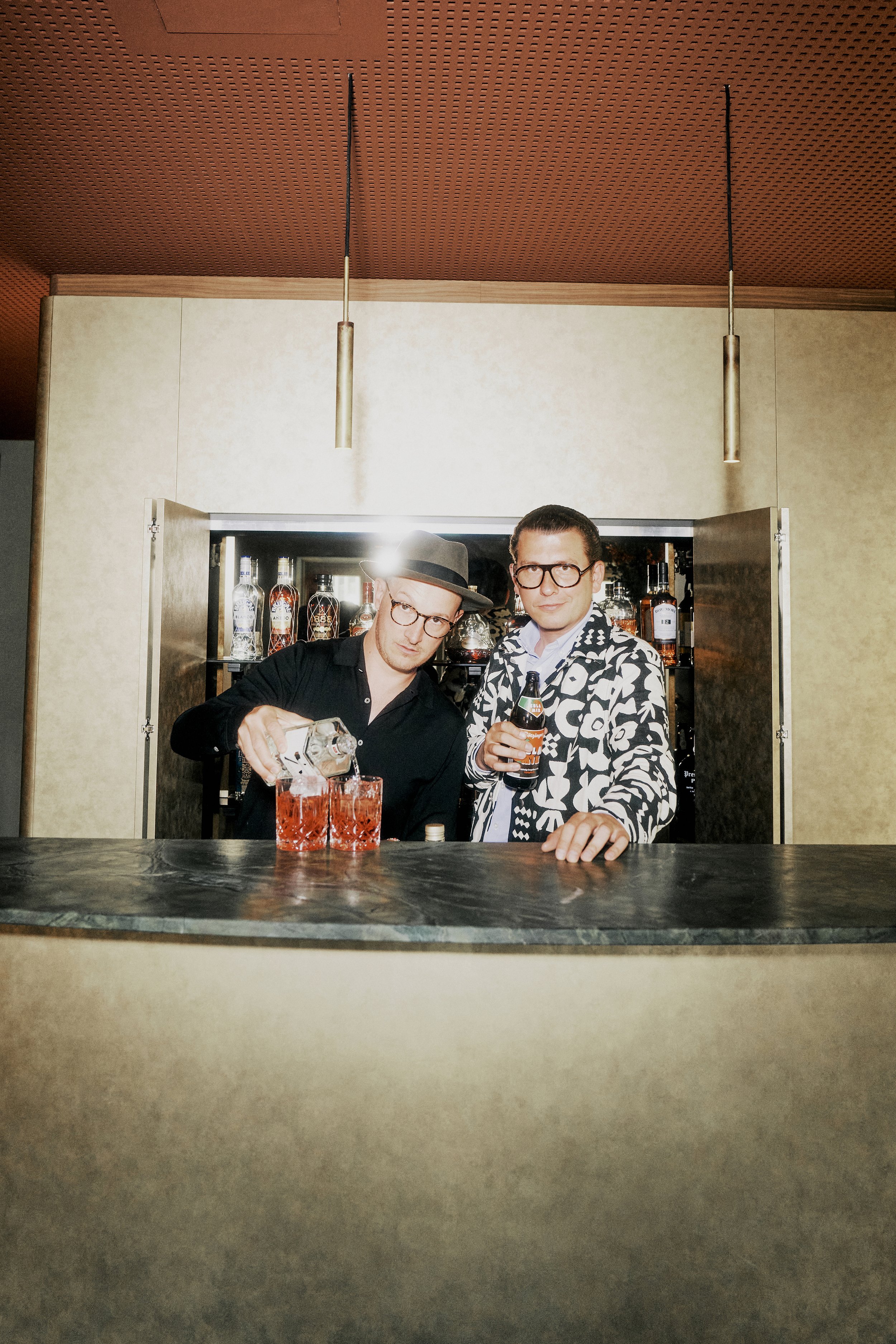 Fabian Freytag und Martin Eggert hinter einer Bar, einer gießt Getränke in Gläser ein. Beide tragen Brillen und sind schick gekleidet. Im Hintergrund eine Auswahl an Spirituosen in einem Regal.