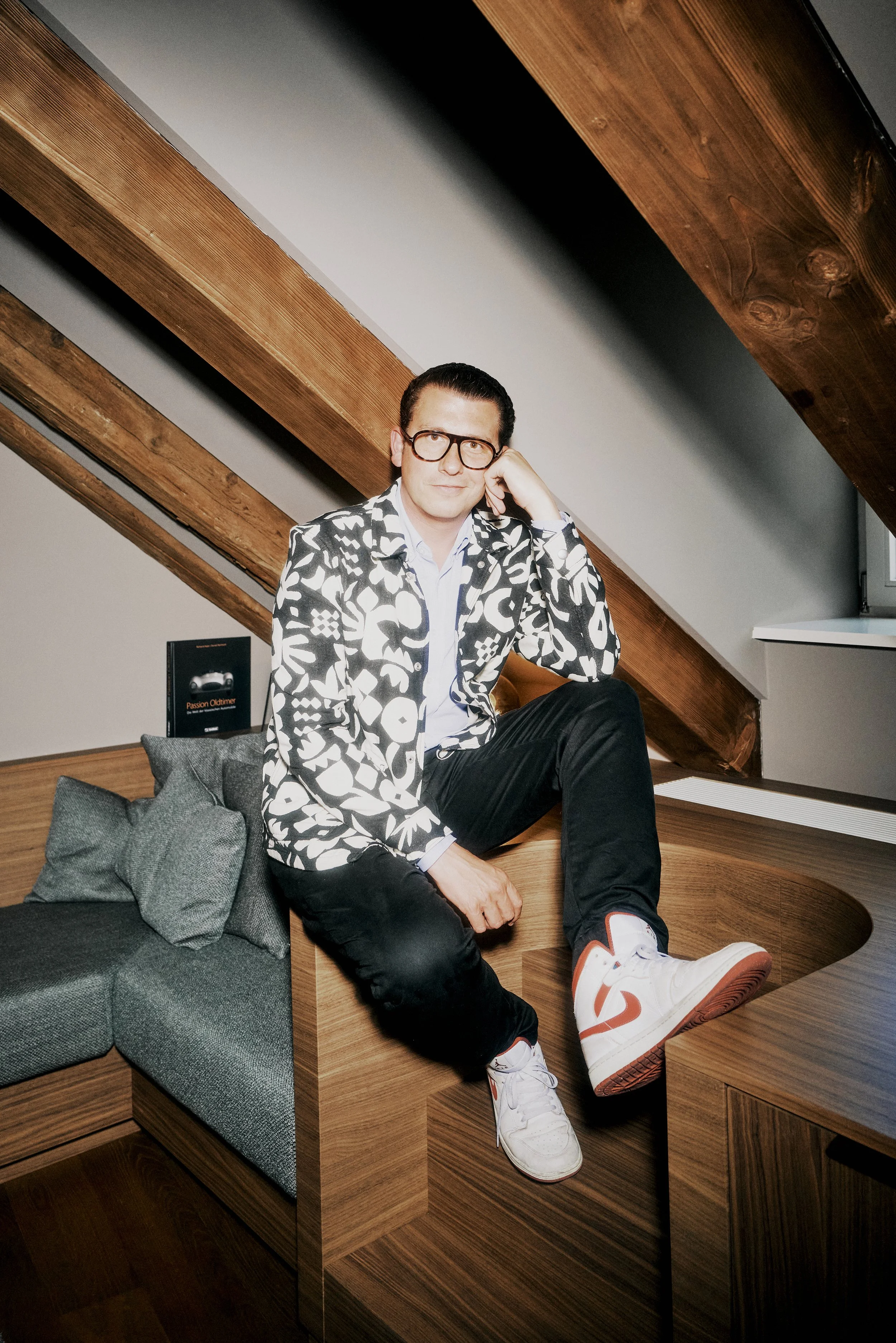 Fabian Freytag mit Brille und gemustertem Jackett sitzt auf einem Holzmöbel unter modernen Dachbalken.