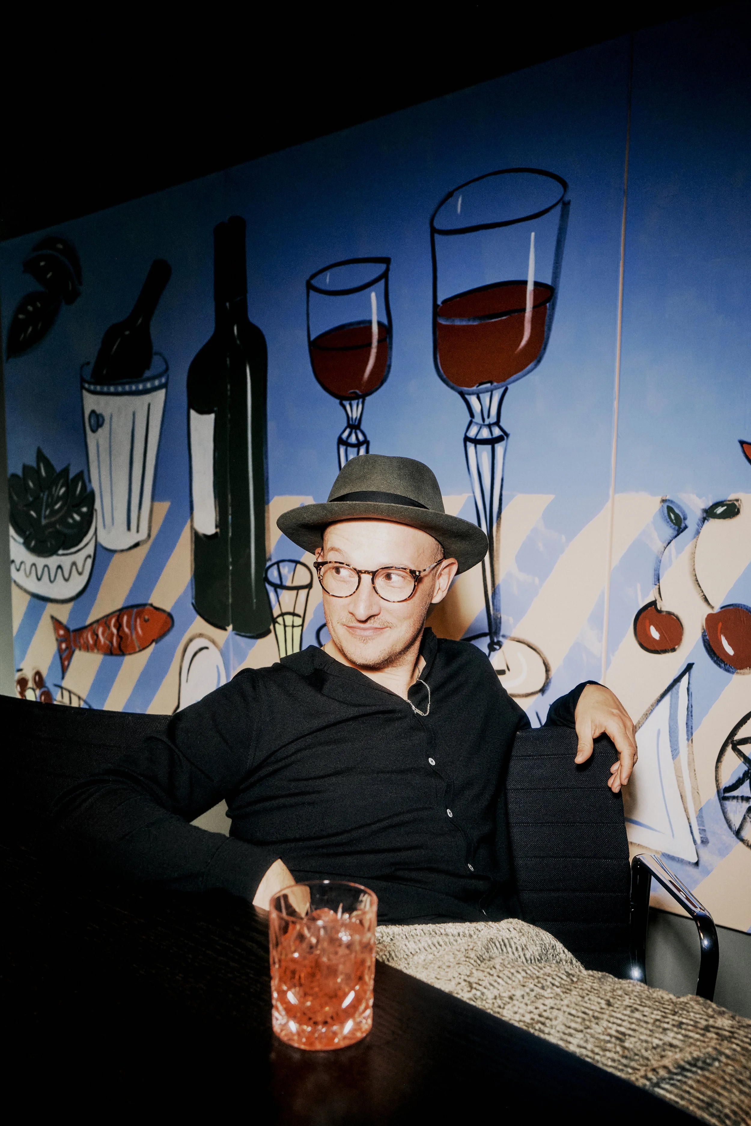 Eine Person mit Hut und Brille sitzt an einem Tisch mit einem Getränk in einem Glas. Im Hintergrund ist ein Wandgemälde mit Weinflaschen und Gläsern zu sehen.