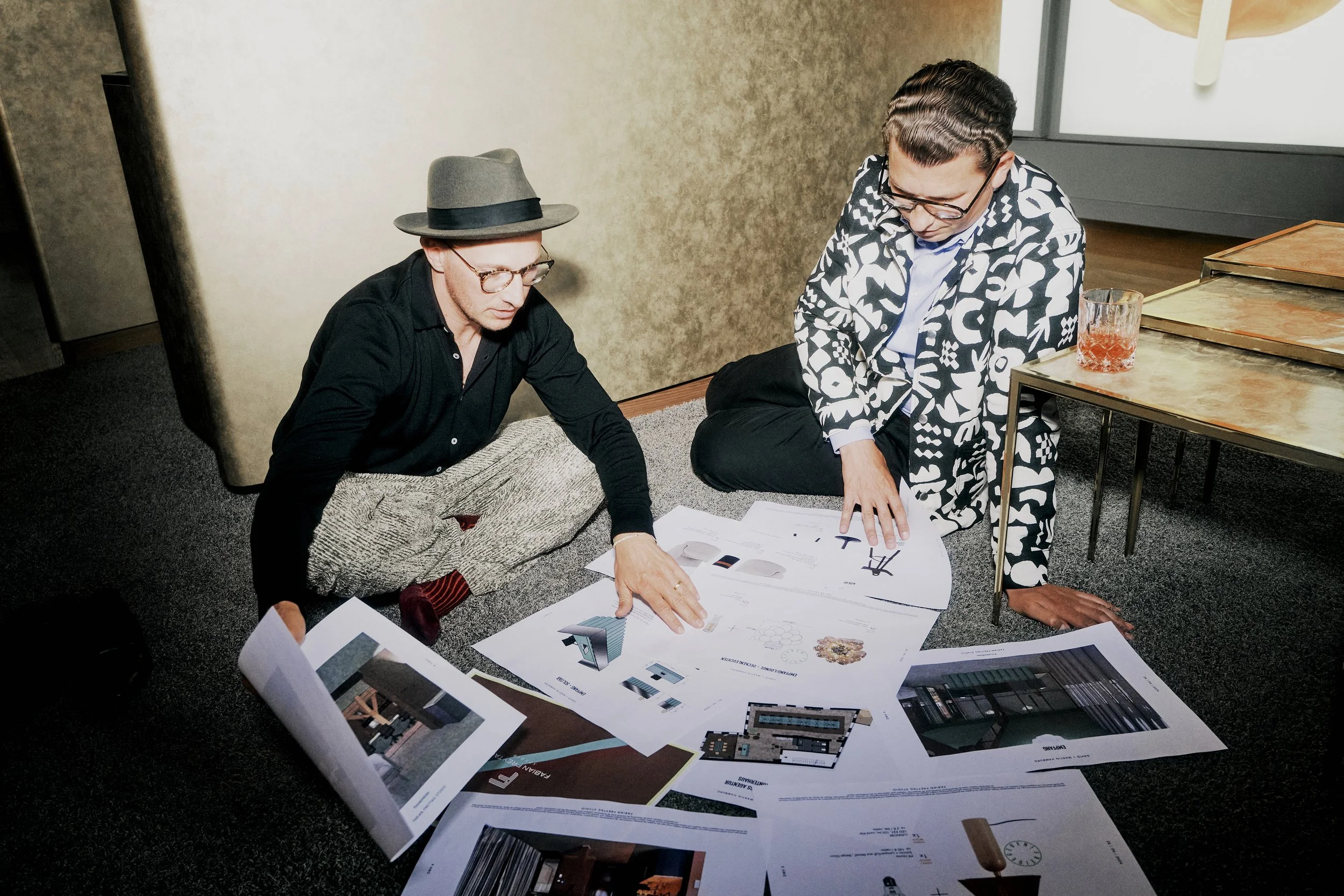 Zwei Männer sitzen auf dem Boden und betrachten Architekturzeichnungen, umgeben von mehreren Blättern. Einer trägt einen Hut, der andere eine gemusterte Jacke. Ein Getränk steht auf einem Tisch neben ihnen.