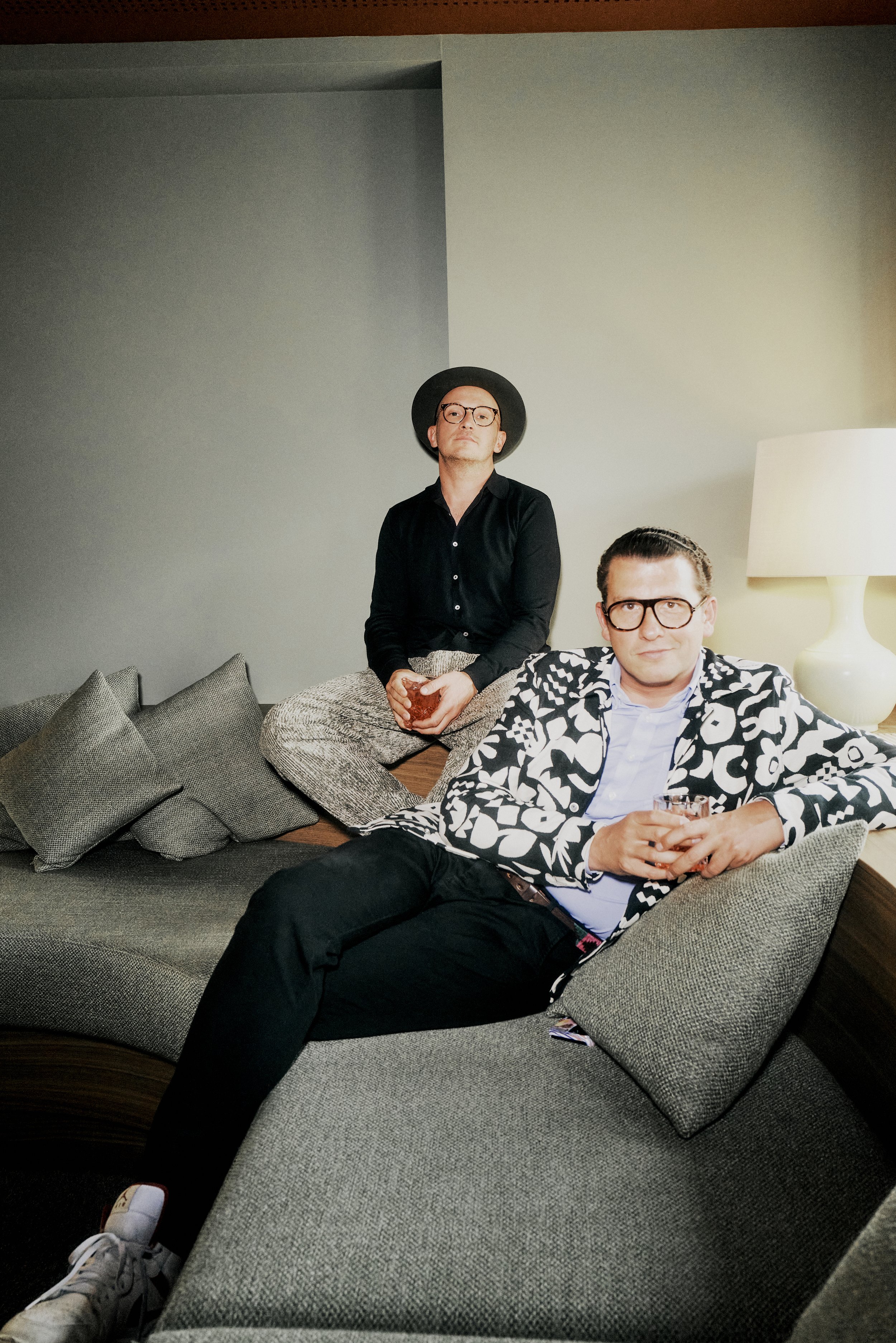 Fabian Freytag und Martin Eggert sitzen auf einem modernen, grauen Sofa. Der Mann hinten trägt einen schwarzen Hut und eine Brille, der andere Mann trägt ein auffälliges, weiß-schwarz gemustertes Sakko. Im Hintergrund ist eine Lampe zu sehen.