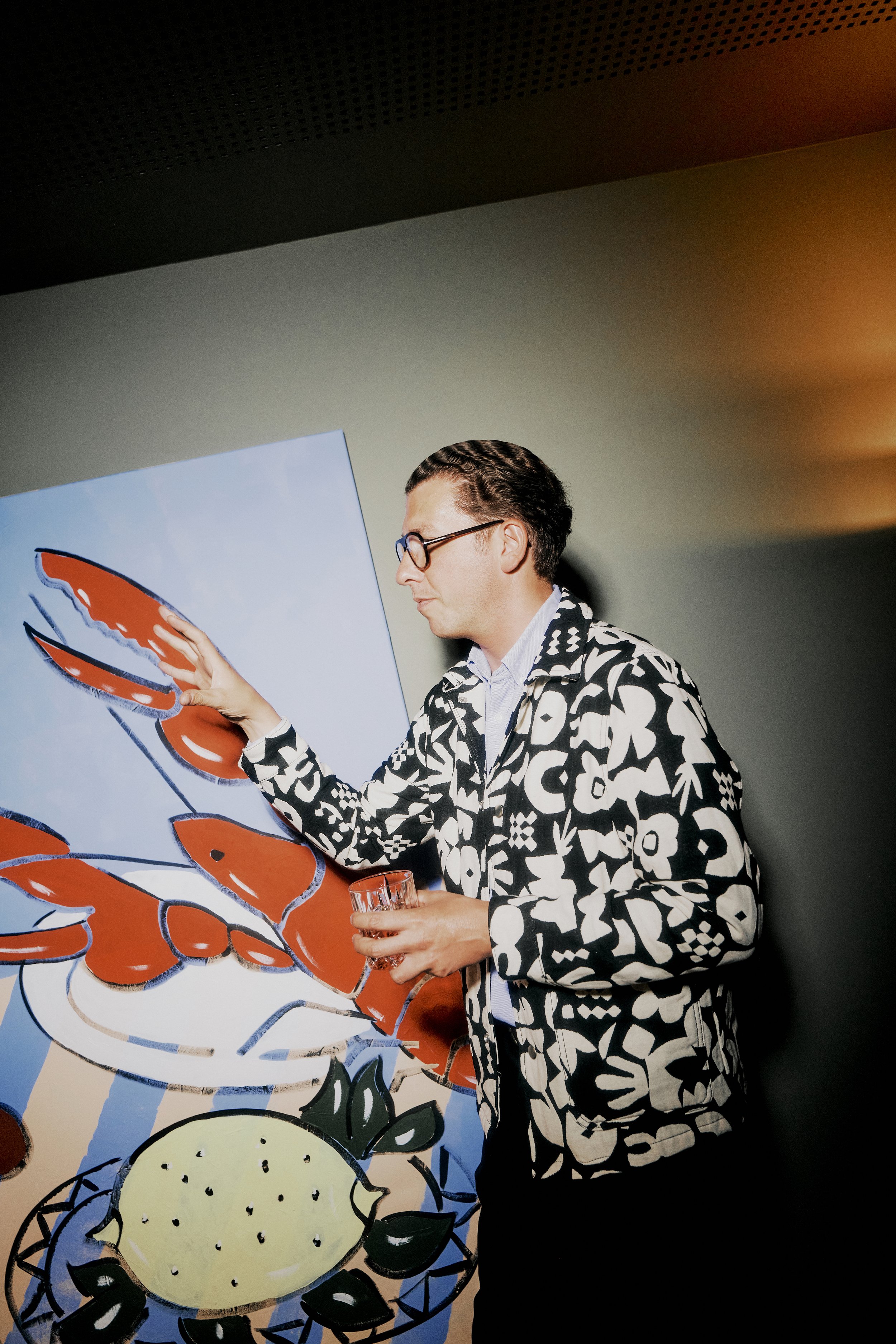 Ein Mann mit Brille und einem gemusterten Jackett steht vor einem Gemälde mit einem Hummermotiv und einer Zitronenscheibe, hält ein Glas in der Hand.