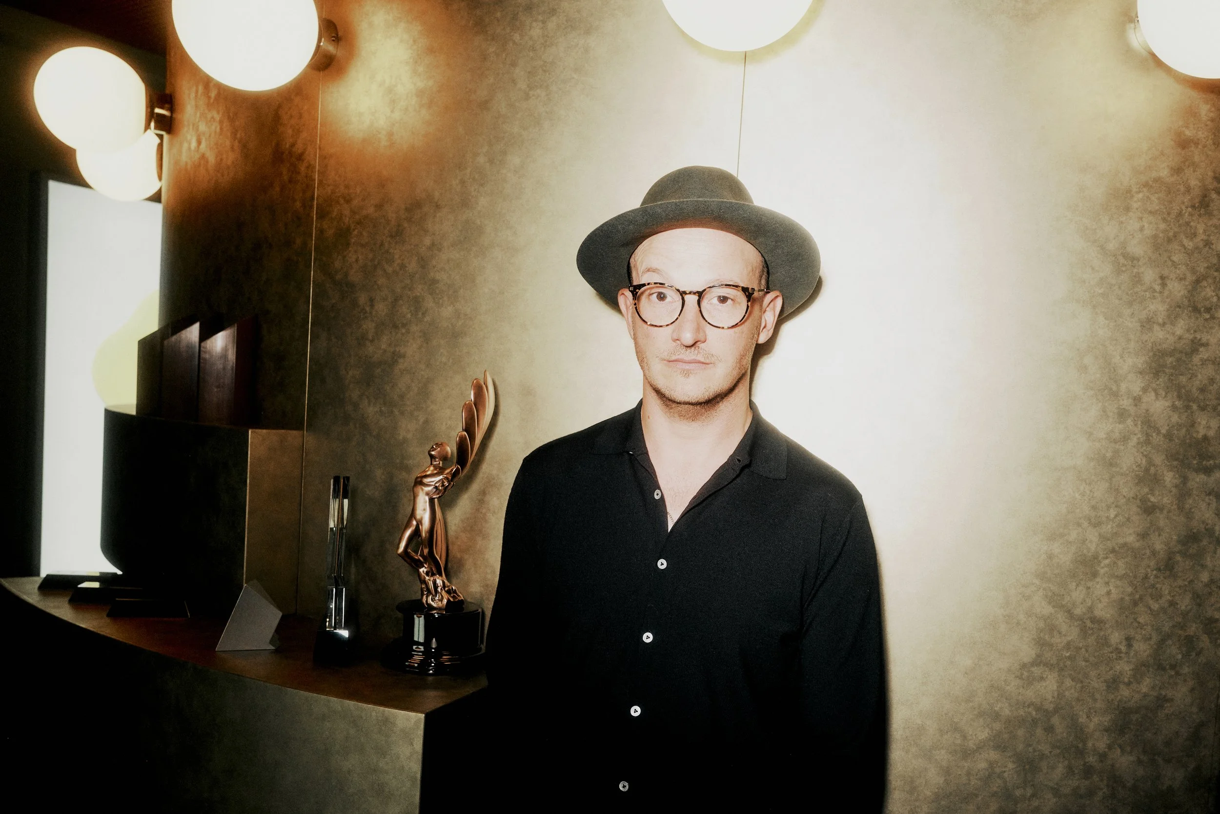 Ein Mann mit Brille und Hut steht in einem Raum mit kupferfarbenen Wänden. Neben ihm auf einem Tisch befinden sich mehrere Awards und Trophäen. Die Beleuchtung ist warm und es sind runde Wandlampen zu sehen.