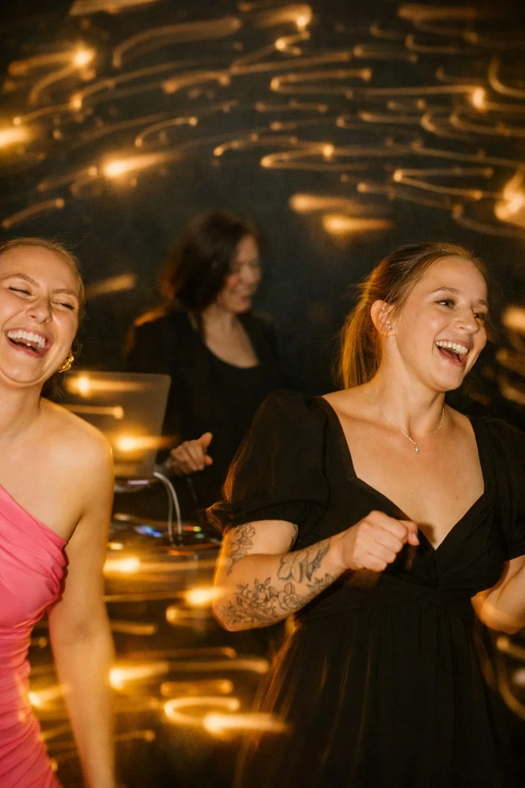 Two women are laughing and dancing at a party with a DJ in the background. The scene has streaks of light indicating movement and lively energy.