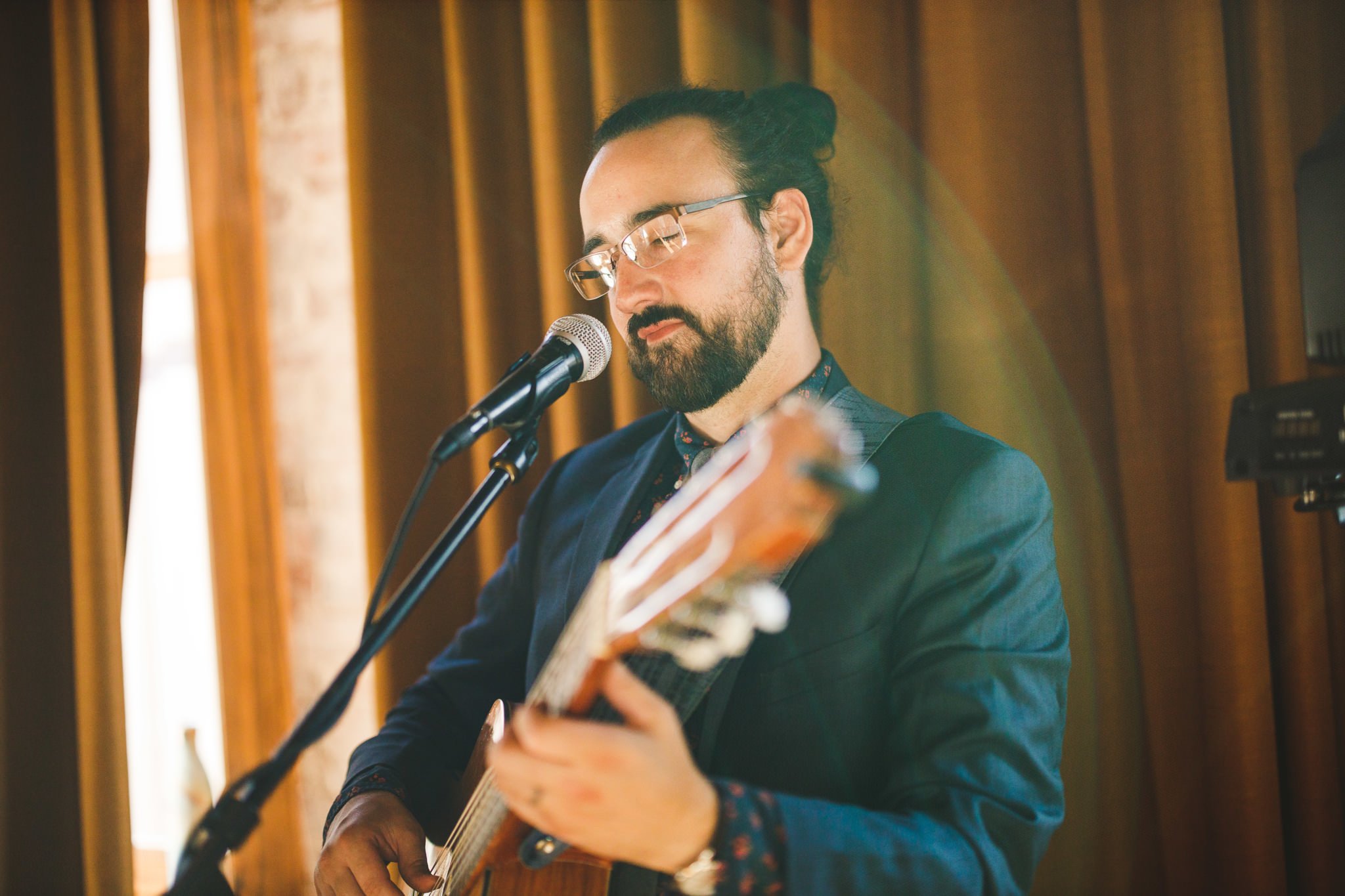 A man with glasses and a beard, wearing a dark blazer, is playing an acoustic guitar in front of a microphone in a wooden-paneled room.