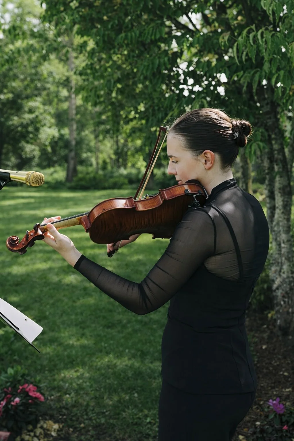 A woman with dark hair tied in a bun is playing a violin outdoors in a park-like setting with green trees and grass.