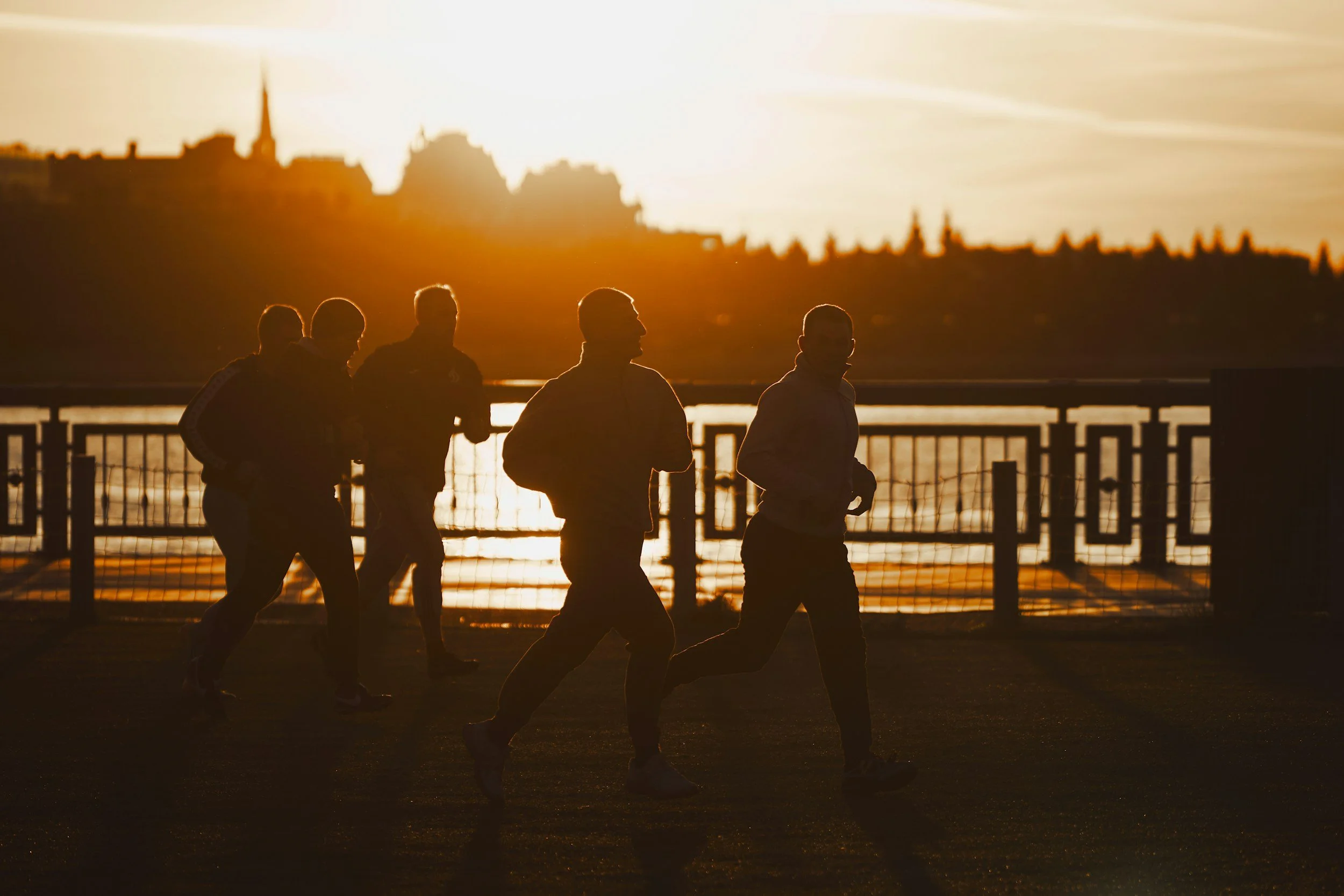 Fünf Menschen joggen am Flussufer während des Sonnenuntergangs, die Silhouetten sind sichtbar vor der warmen Sonne am Himmel.