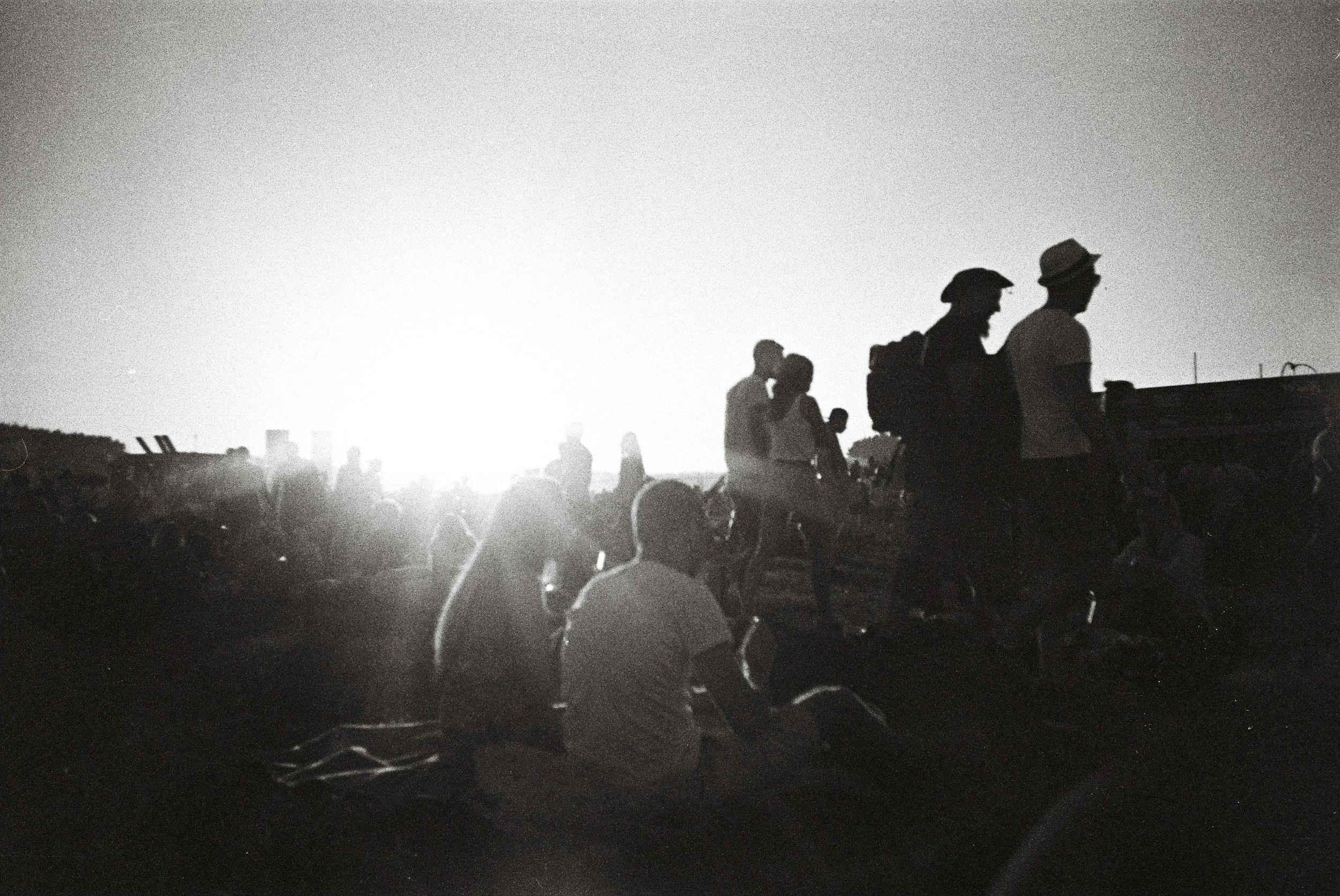 Schwarz-weiße Fotografie einer Menschenmenge im Sonnenuntergang, einige sitzen, andere stehen oder gehen.