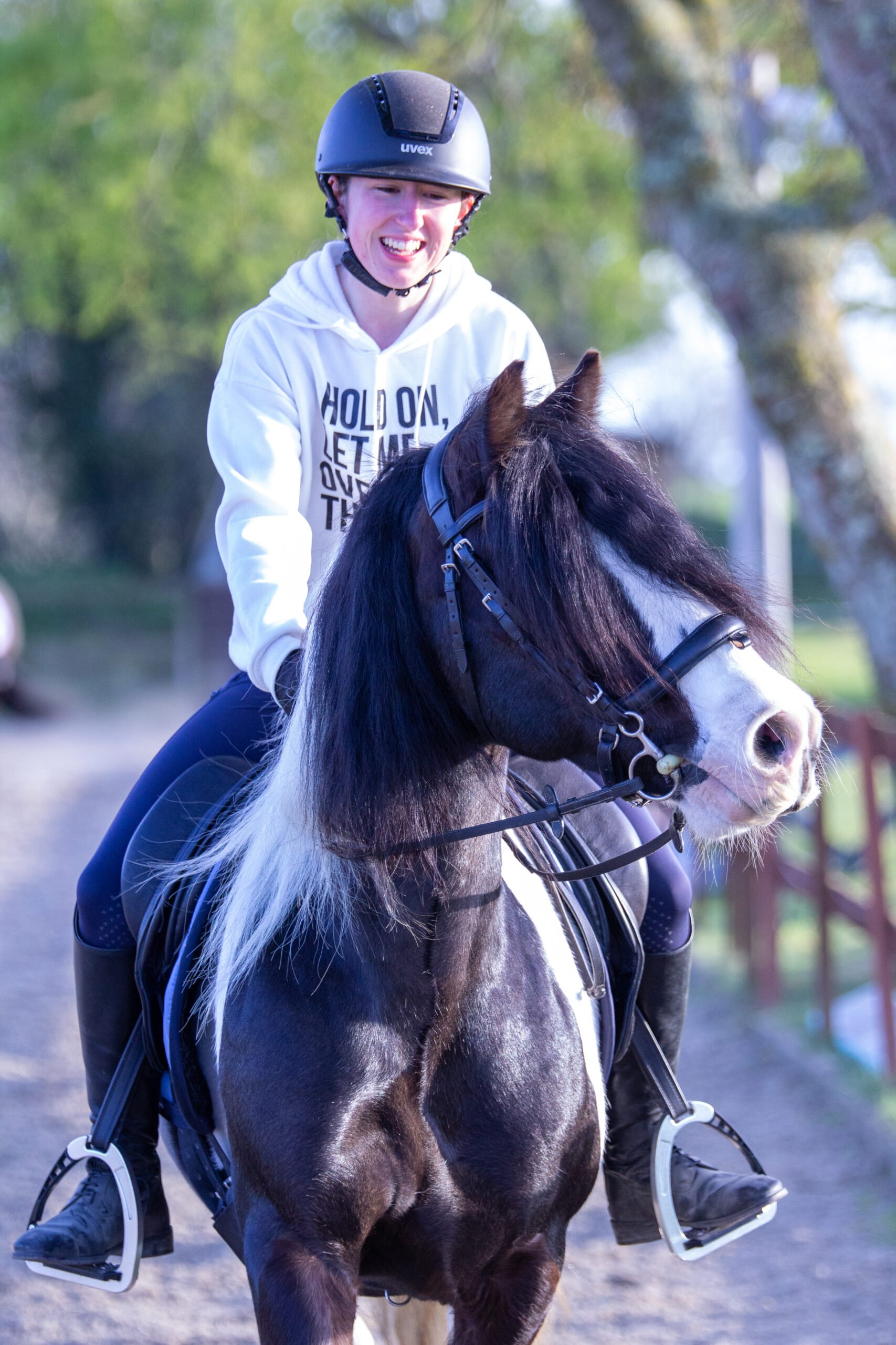 A young man smiling while riding a black and white horse outdoors with trees in the background.