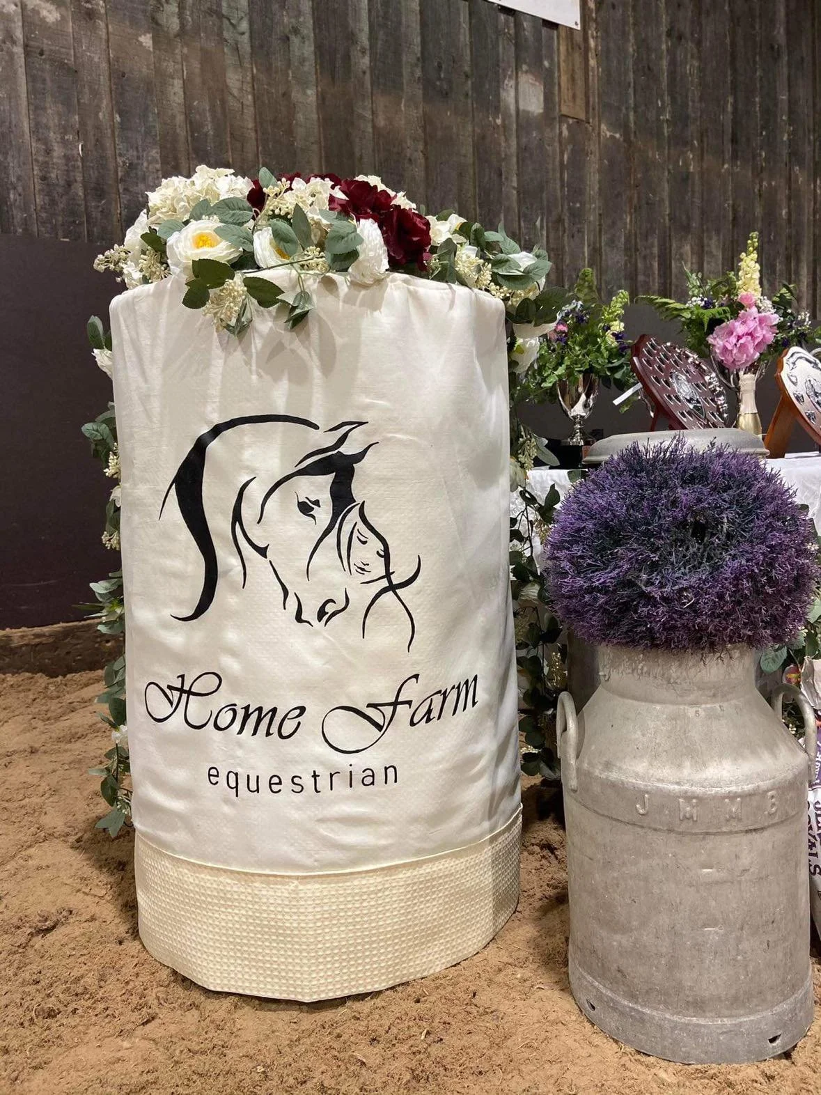 A decorated area with a white fabric cover featuring a horse drawing and the words 'Home Farm Equestrian', adorned with flowers and greenery, with a large purple flowering plant in a white cement container nearby, and a wooden fence in the background.