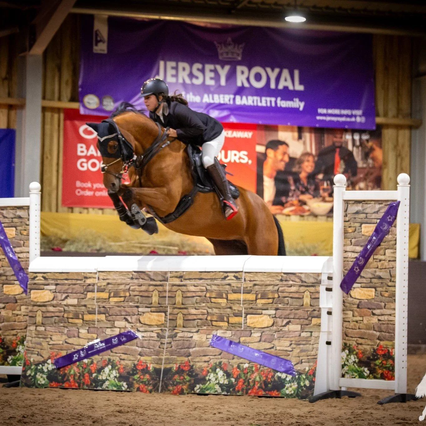 A woman riding a horse jumping over an obstacle in an indoor equestrian arena with banners and advertisements in the background.